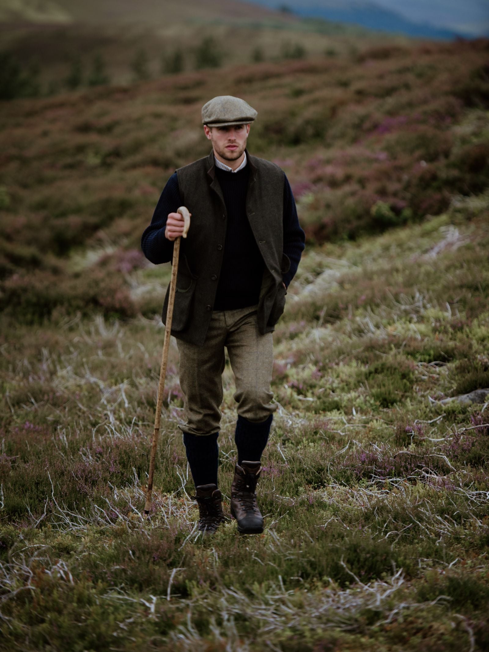 A man wearing Campbells of Beauly Plus Twos and a classic flat cap strolls with a wooden walking stick across a softly colored, heather-covered hillside. The natural landscape complements his traditional, tailored style.