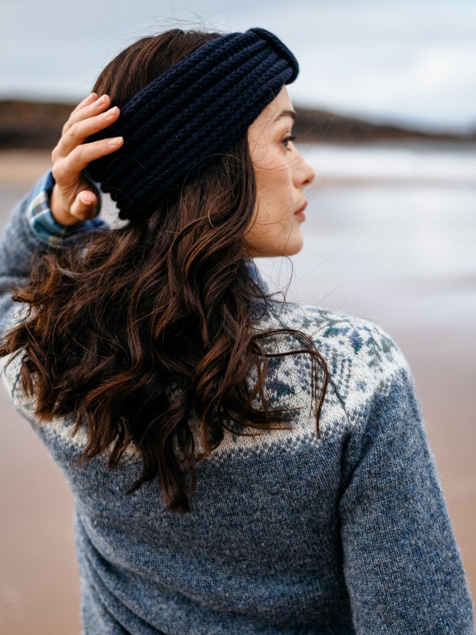 A woman with wavy brown hair wears a Campbell's of Beauly Cashmere Ear Warmer and a blue patterned sweater, standing on a sandy beach and facing away as she touches her head. The blurred background shows water and sky.