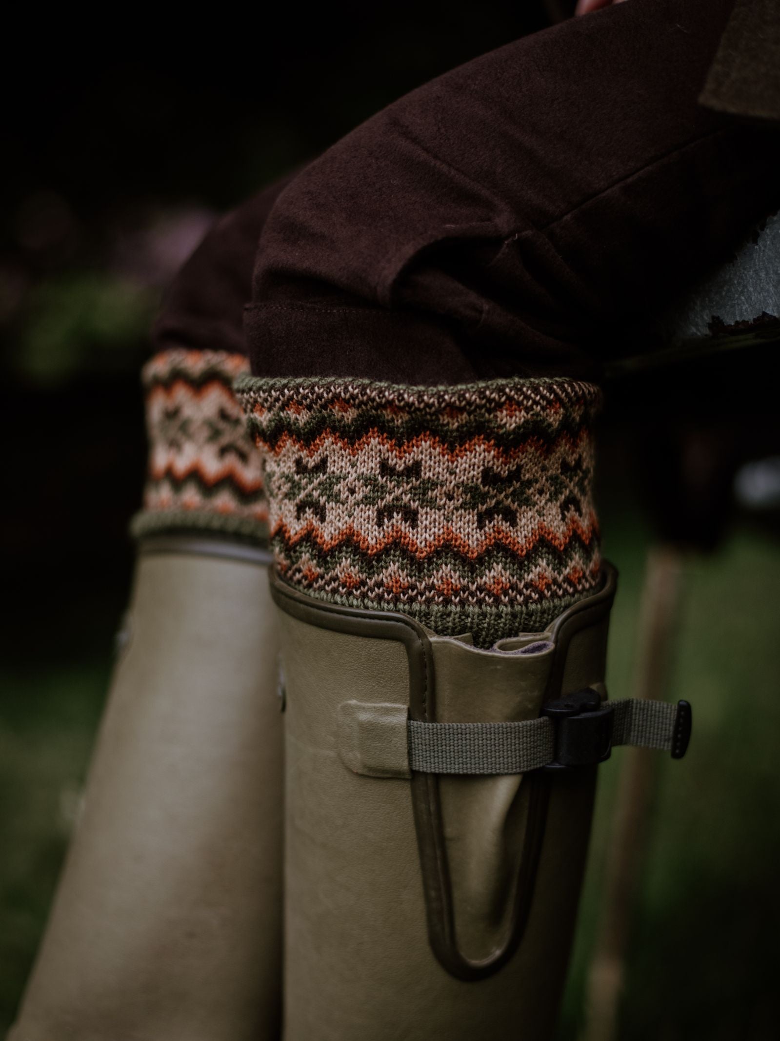 A close-up shows Campbells of Beauly Merino Fairisle Shooting Socks—with an orange and green pattern—visible above olive green rubber boots and brown pants, set against blurred greenery to highlight the stylish knitwear.