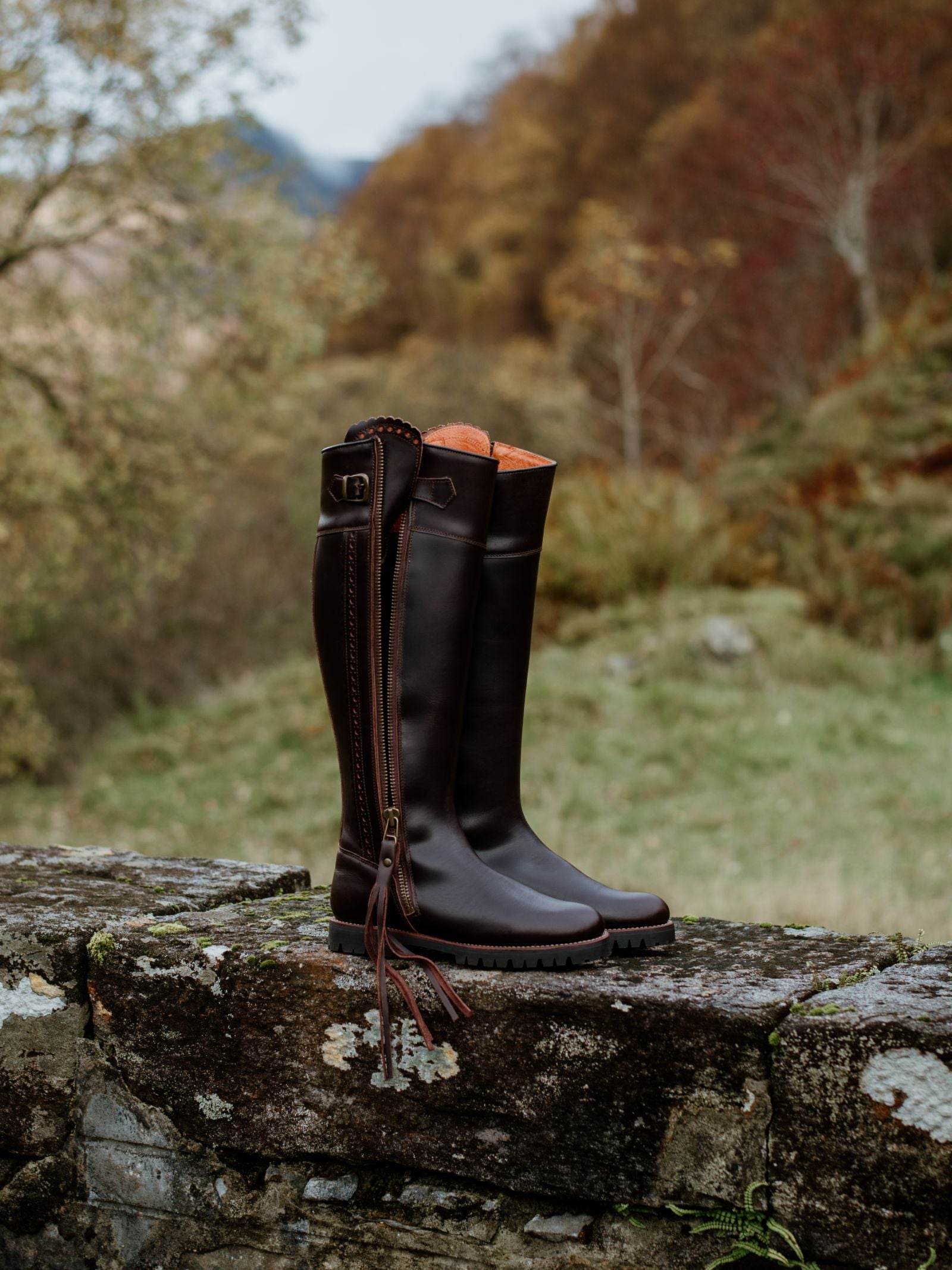 A pair of tall, dark brown Campbells of Beauly Leather Spanish Boots with side zippers, tassels, and a water-resistant breathable membrane sit on a stone wall against blurred autumn trees and rolling hills.