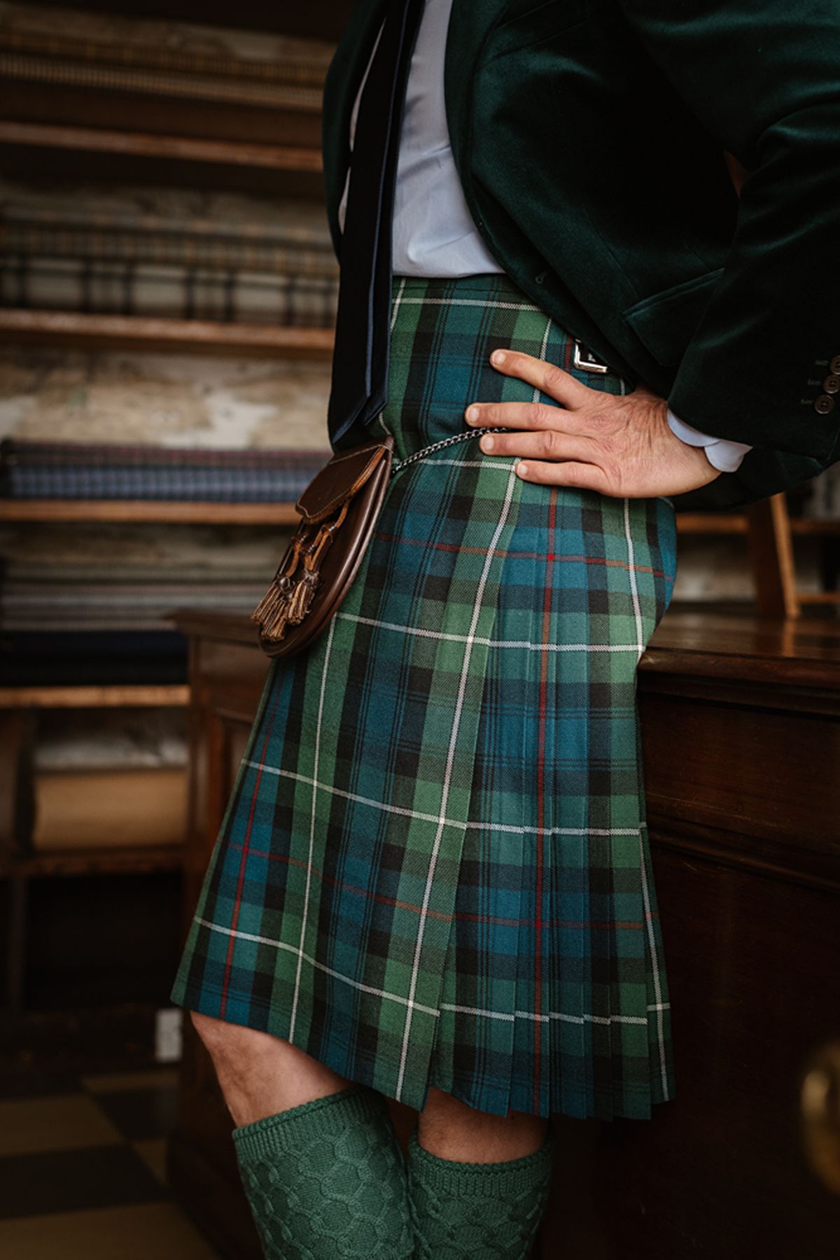 A person wearing a green tartan kilt, a dark jacket, and a sporran, leaning against a wooden counter.