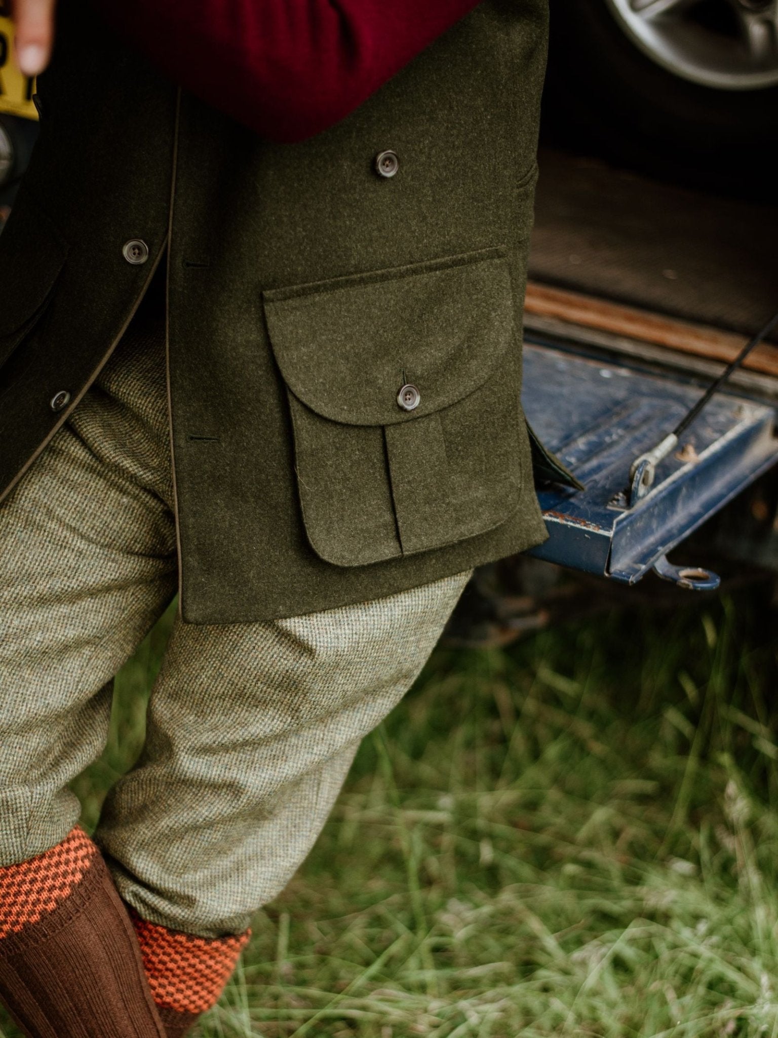 A person wearing the Campbell's of Beauly Luxury Nehru Gilet in Loden green, tan pants, and red-orange knee-high socks sits on the edge of a vehicle’s open trunk with grass below.