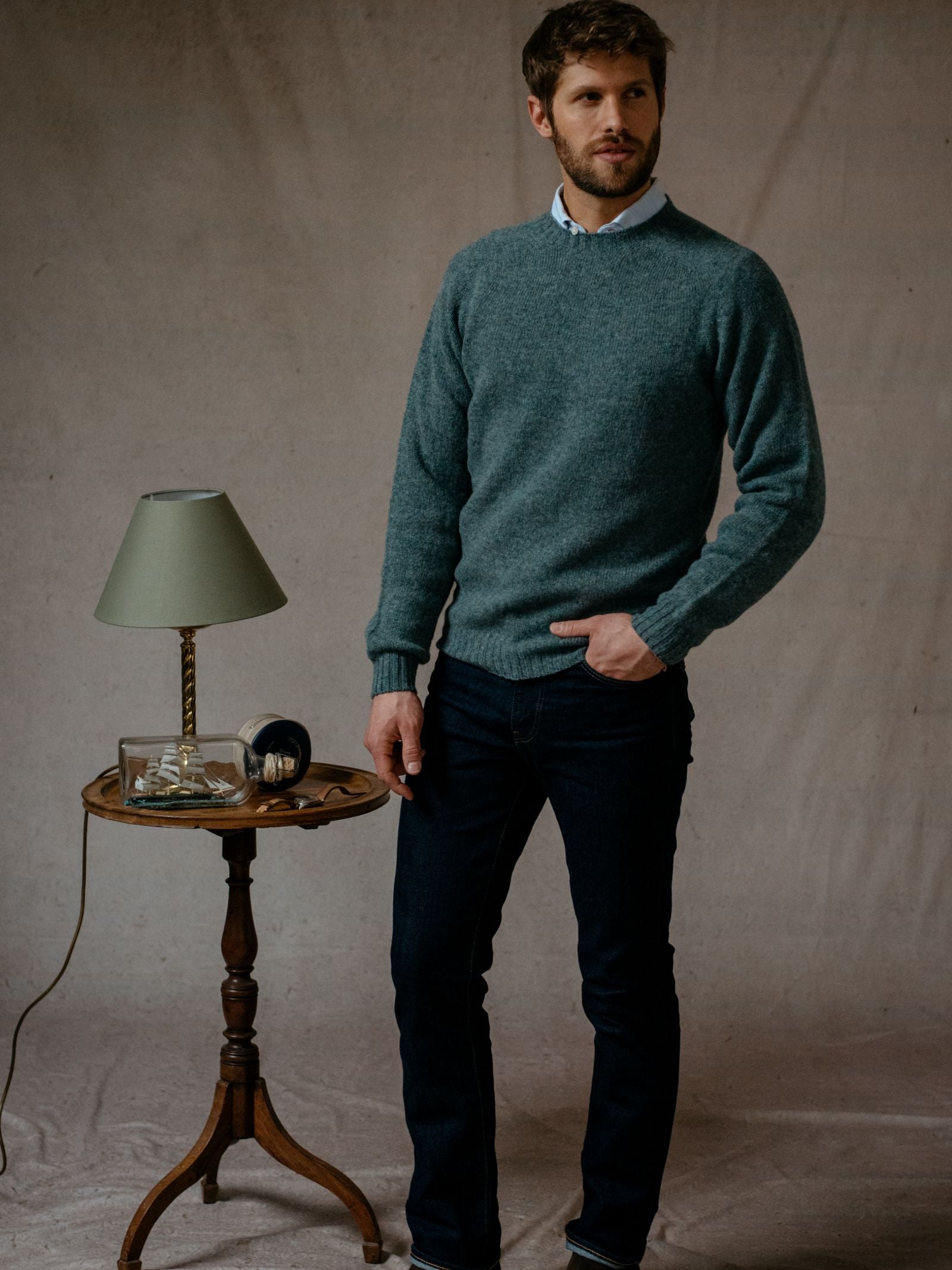 A man wears a Campbells of Beauly Shetland Jumper with dark jeans, standing by a small round wooden table topped with a green lampshade, glass dish, and black alarm clock against a plain beige backdrop.