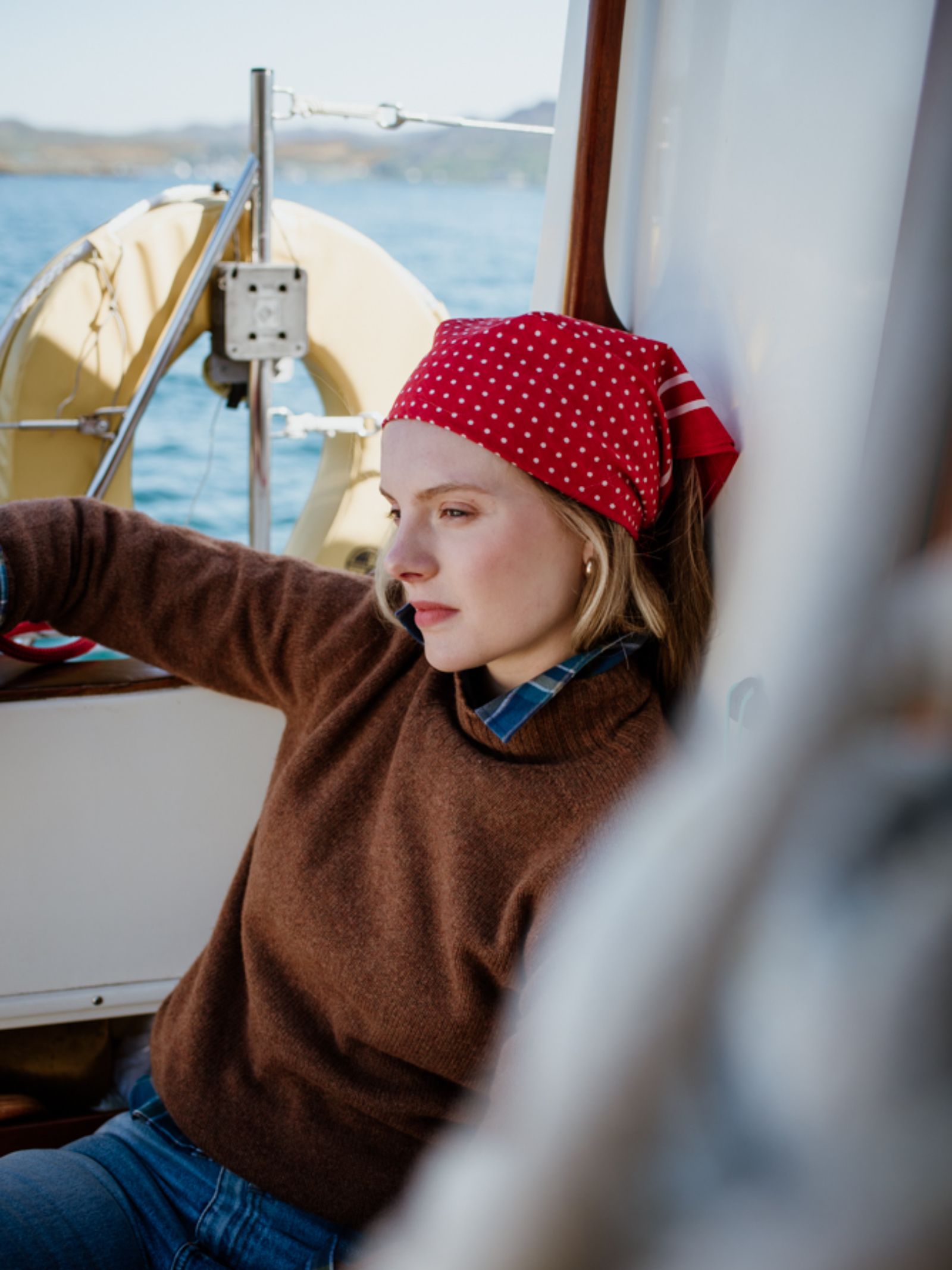A woman wearing the Campbells of Beauly Cotton Spotty Hank handkerchief and brown sweater sits thoughtfully on a boat, with a life ring and water behind her, highlighting the classic polka dot design in various colours.