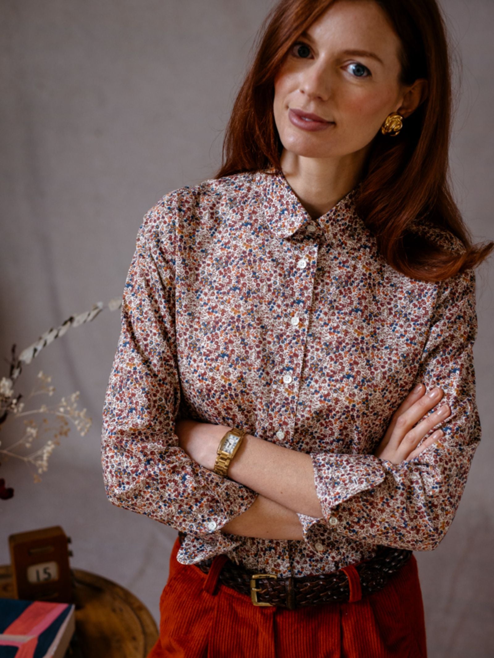 A woman with long red hair wears a Campbells of Beauly Liberty Print Cotton Shirt, red corduroy pants, and gold earrings. She stands with arms crossed, looking at the camera. In the background is a small table with books and dried flowers.