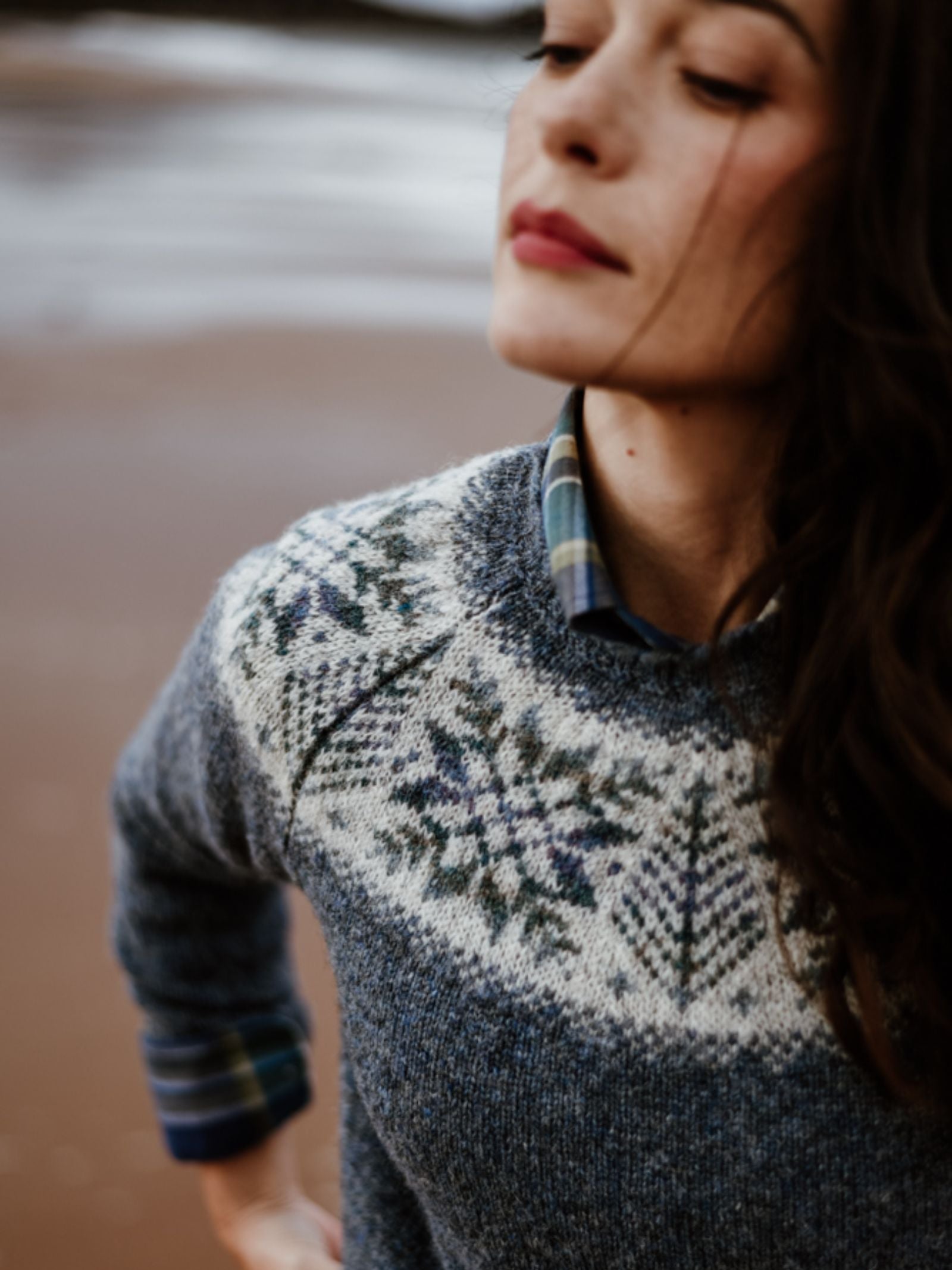 A woman with long brown hair stands outdoors by sand and water, serenely wearing Campbell's of Beauly Shetland Wool Snowflake Crew Jumper in blue and white over a plaid shirt.