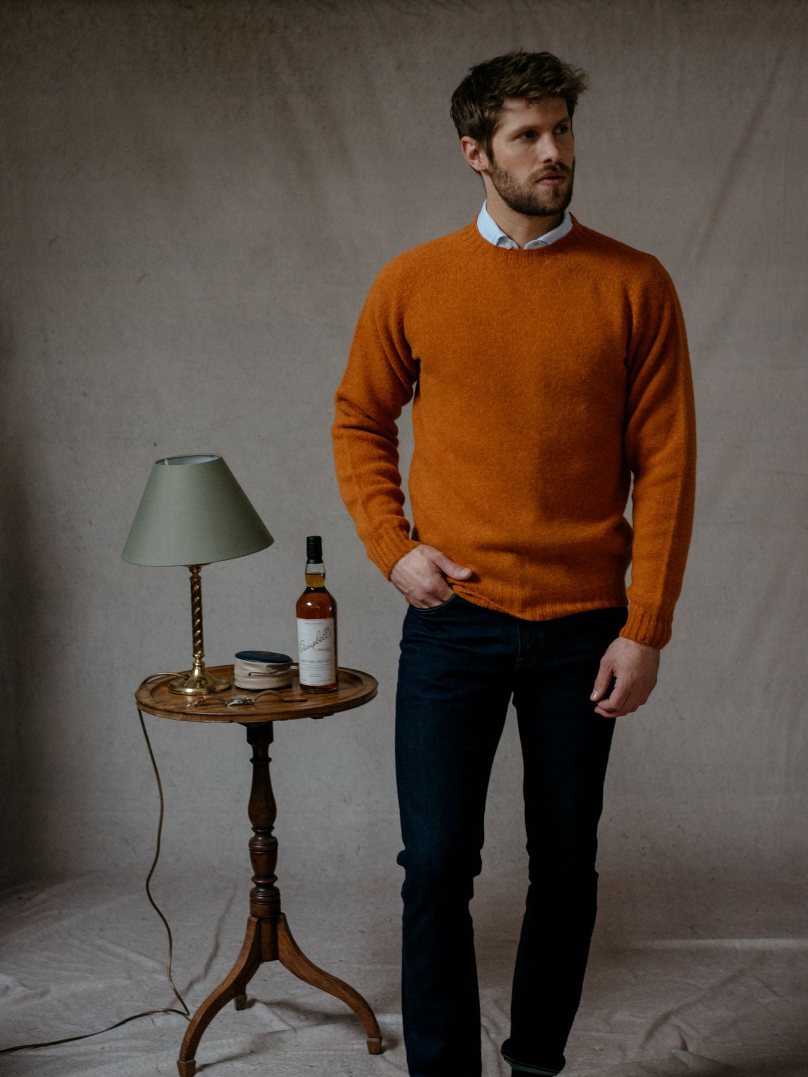 A man in a Campbells of Beauly Shetland Jumper with dark jeans stands next to a small round table holding a green lamp, a bottle, and a glass, set against a neutral backdrop.