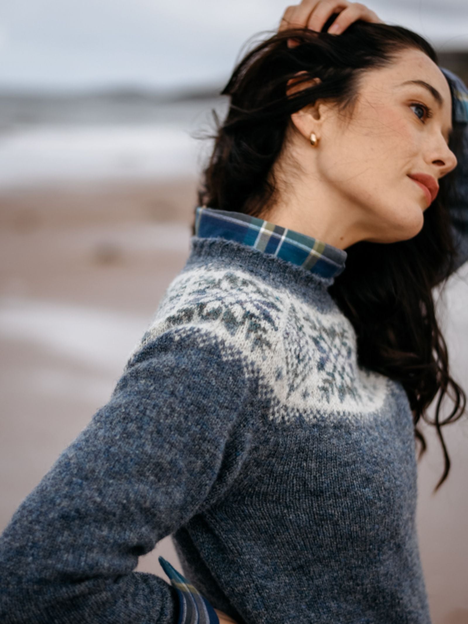 A woman with dark hair wears the Campbell's of Beauly Shetland Wool Snowflake Crew Jumper in a blue and white Fairisle design over a plaid shirt, standing on a beach with one hand in her hair. The background is slightly blurred.
