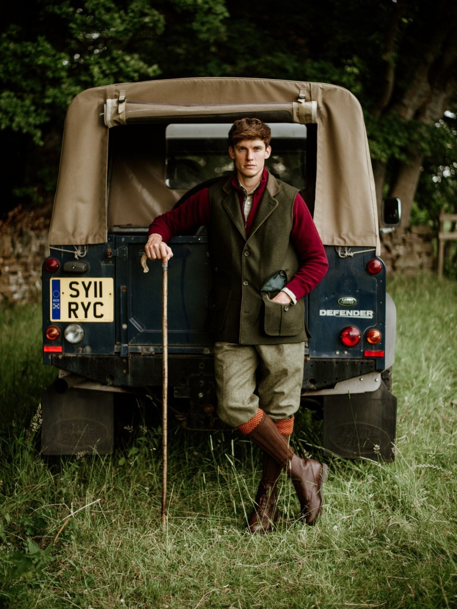 A man in traditional country attire, wearing a Campbells of Beauly Luxury Nehru Gilet in Loden, leans against a Land Rover Defender in a grassy field, holding a walking stick. Trees and a stone wall can be seen behind him.
