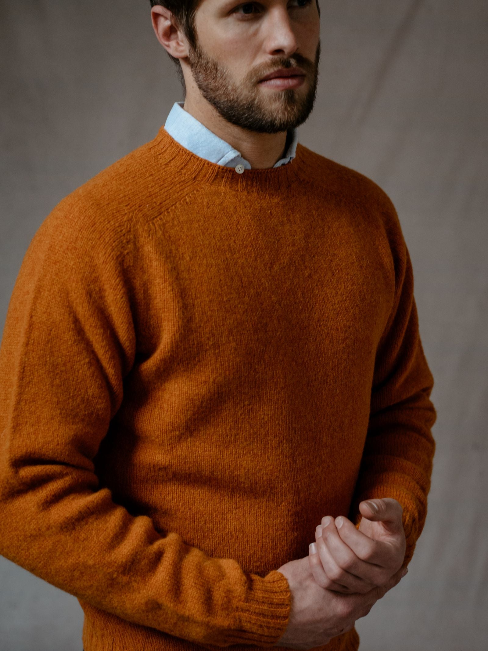 A man with a trimmed beard wears a Campbells of Beauly Shetland Jumper in orange over a light blue collared shirt, standing against a neutral background with his hands gently clasped.