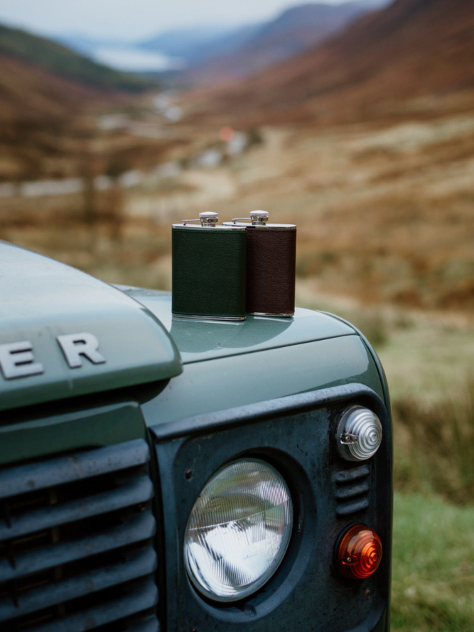 A Campbell's of Beauly x Ettinger 6oz Forest Green Hip Flask rests on a green Land Rover’s hood, set against rugged mountains and misty hills.