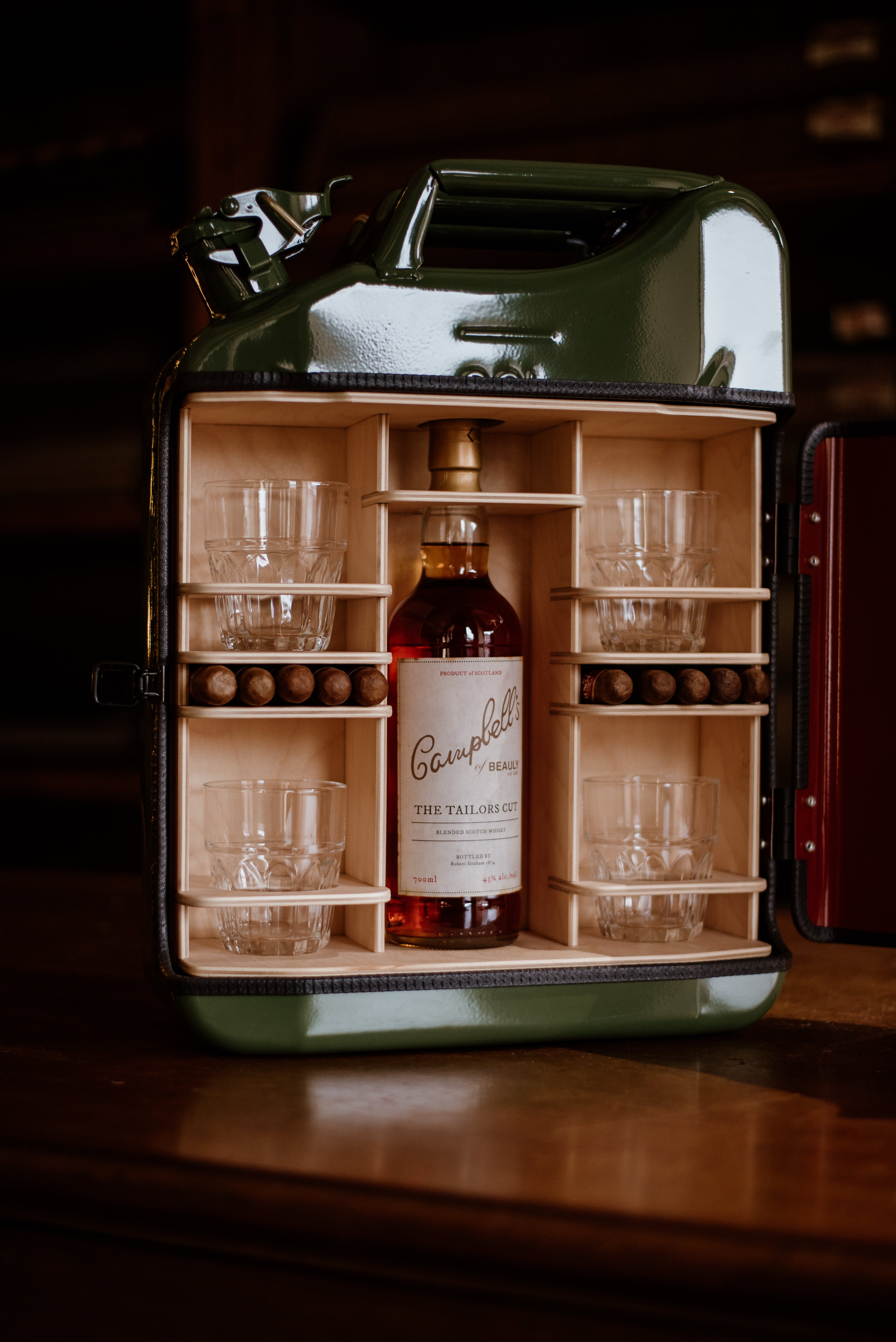 The Connoisseur Upcycled Mini Bar by Campbells of Beauly features a repurposed green jerry can, opened to reveal six glass tumblers, a bottle of Campbell’s whisky, and brown wooden spheres in holders on a wooden base.