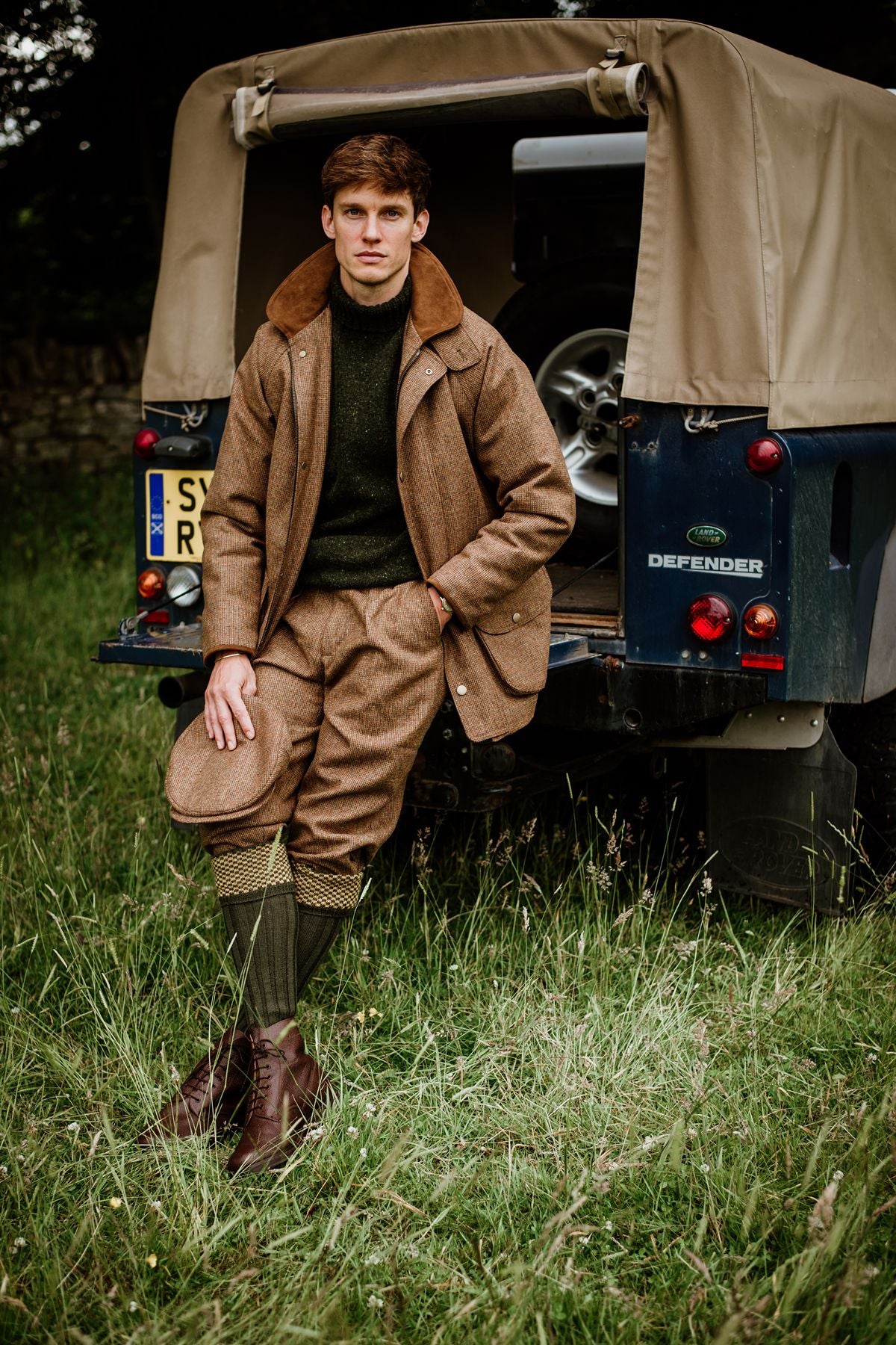 A man in a tweed jacket, plus fours, and a green jumper leans against the back of a blue Land Rover Defender parked in a grassy field.