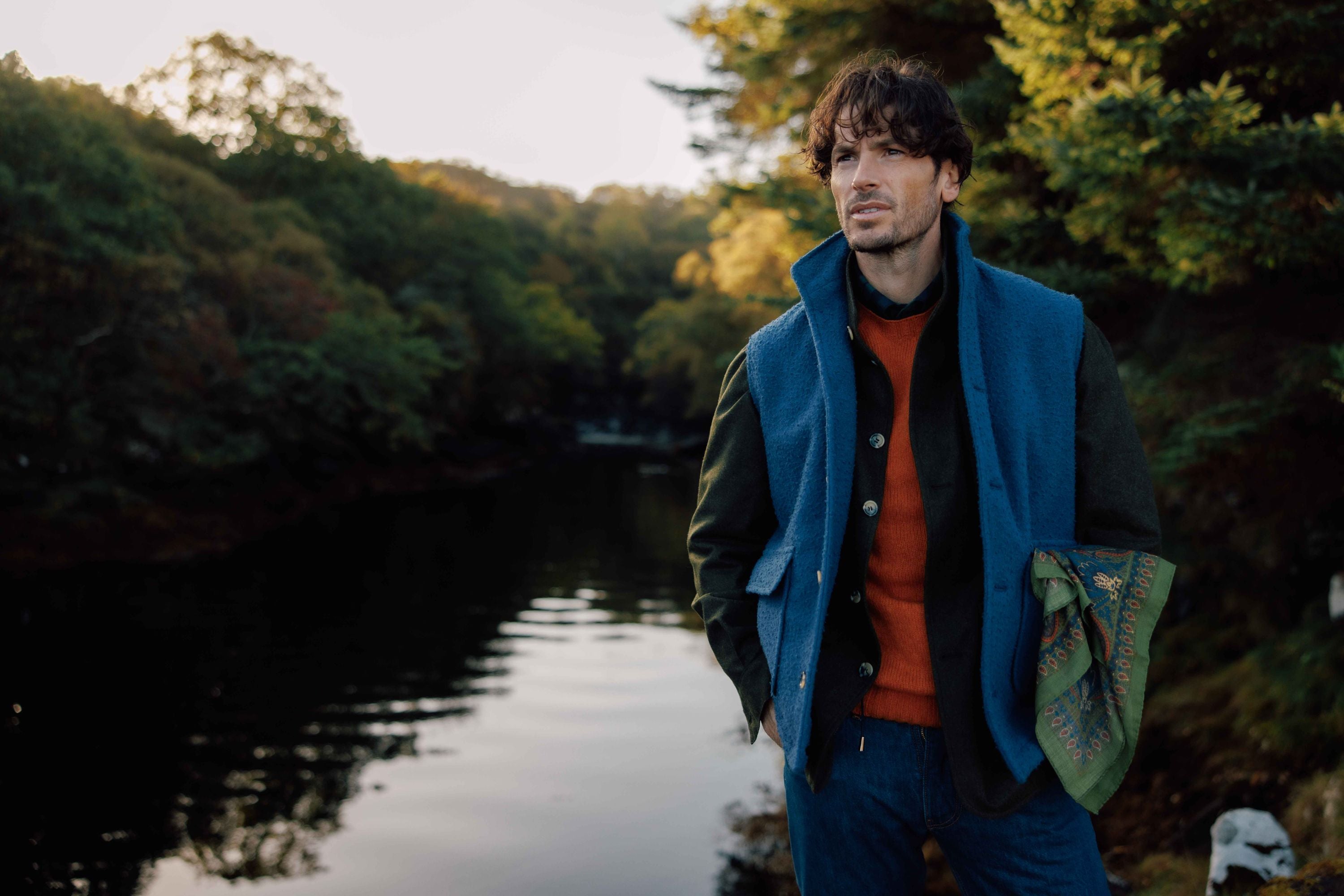 A man stands by a calm river surrounded by trees, wearing a blue vest over a dark jacket and an orange sweater, with a patterned scarf draped over his arm. The background is lush and green, suggesting a peaceful, natural setting.