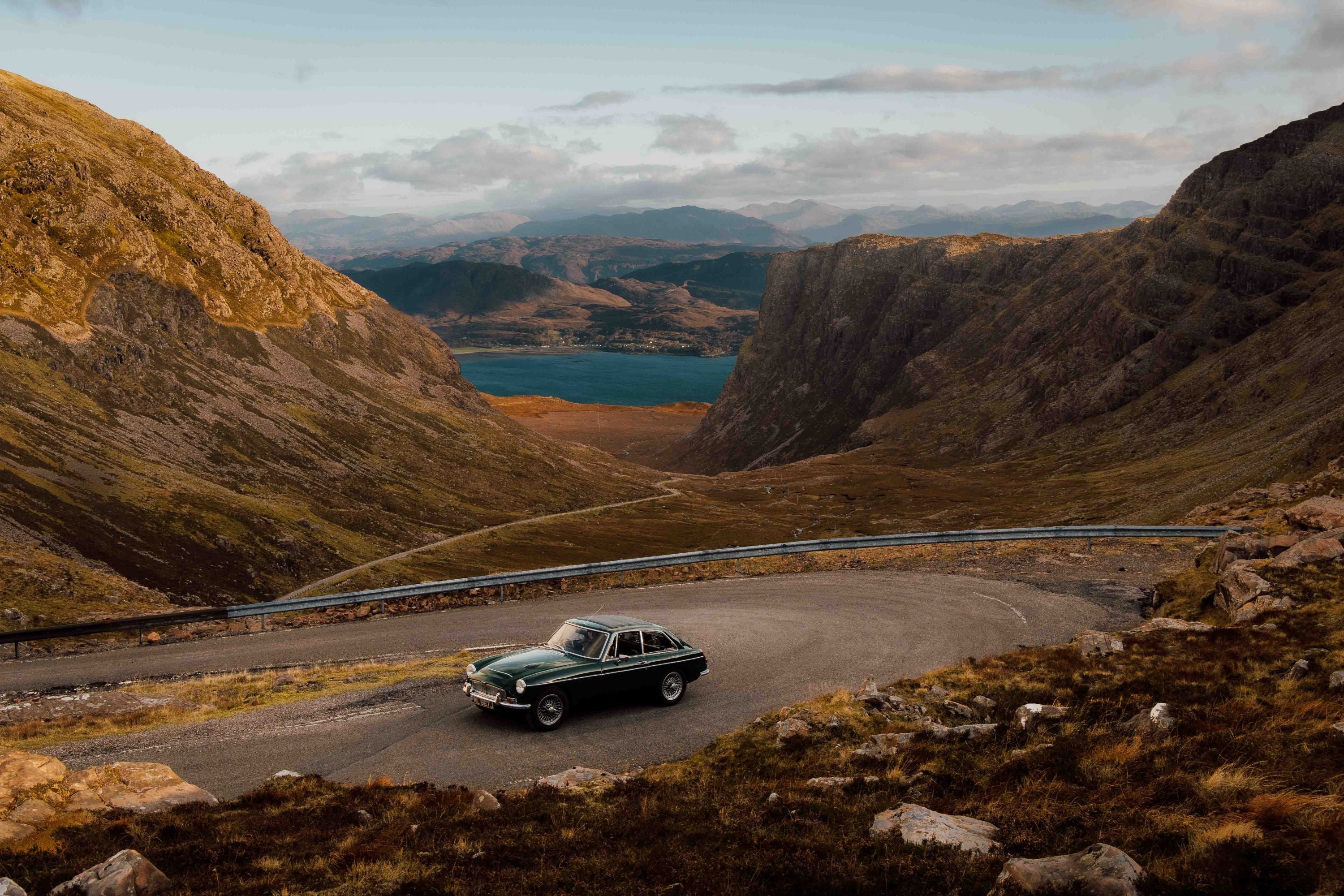 A classic car drives along a winding mountain road surrounded by rocky hills, with a lake and distant mountains visible under a partly cloudy sky.
