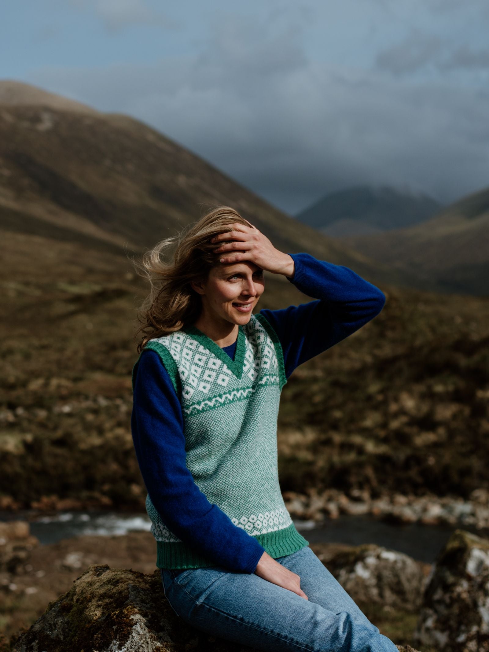 A woman sits on a rock in the mountains, smiling with her hand to her head. She wears Campbells of Beaulys blue and green patterned Nordic V Tank and jeans, with cloudy skies and distant hills behind her.
