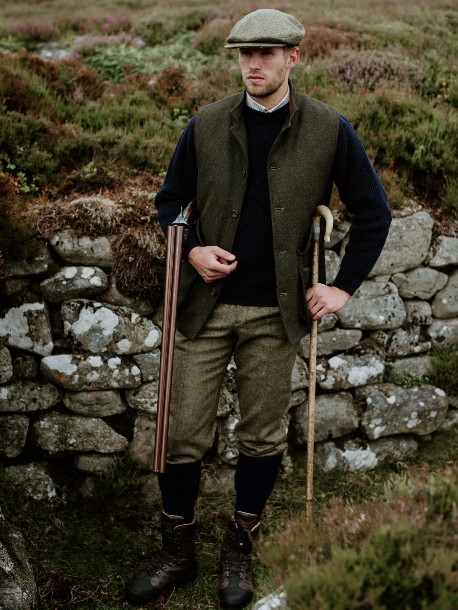 Wearing Campbells of Beauly Plus Twos, a man in classic sporting attire, boots, and flat cap stands by a stone wall outdoors with a walking stick and shotgun, surrounded by green heather and shrubs.