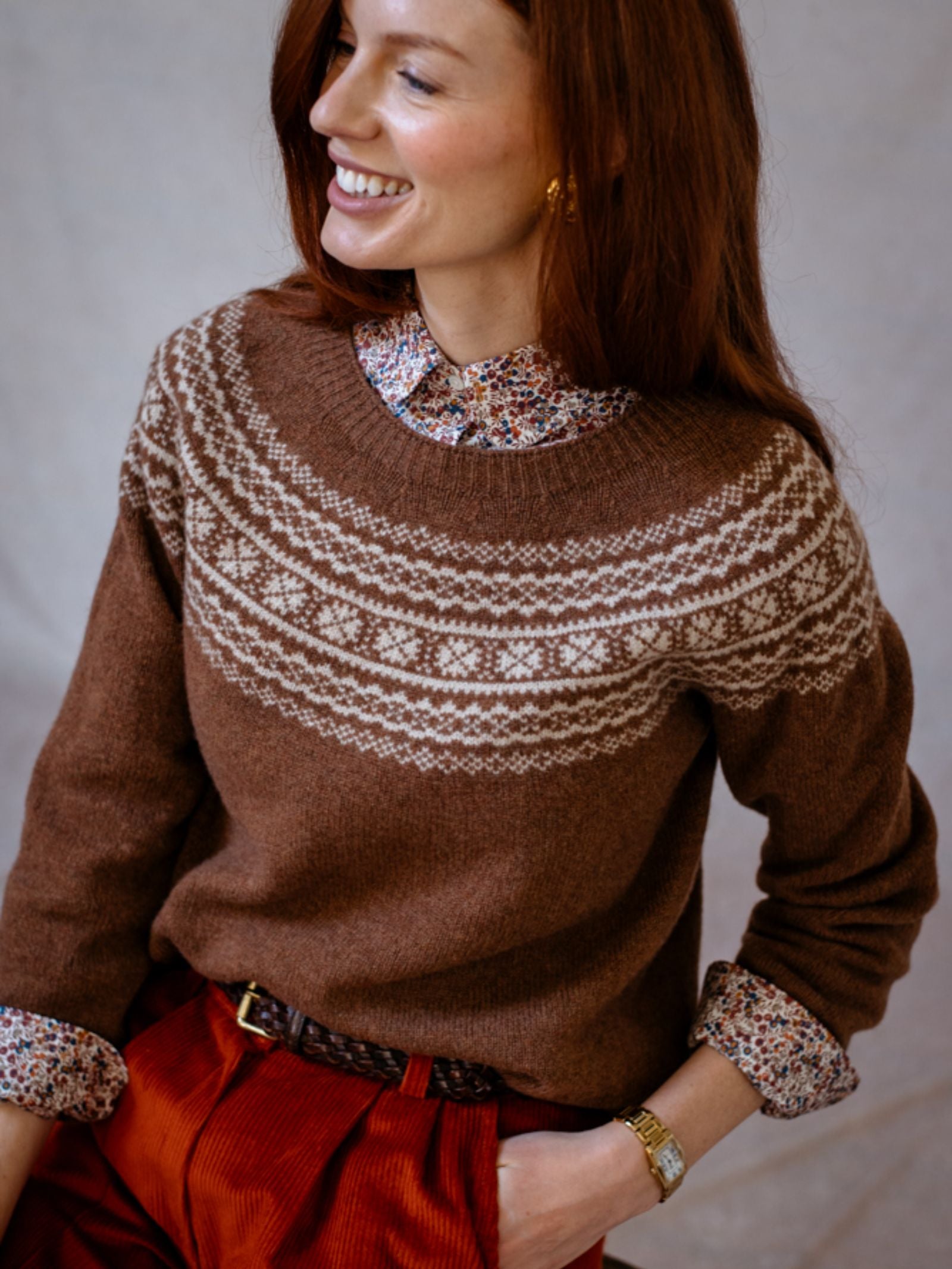 A woman with long red hair smiles, wearing a brown Campbells of Beauly Two-Colour Fairisle Yoke Jumper over a floral-collared shirt and red corduroy pants. She stands with one hand in her pocket against a neutral background.