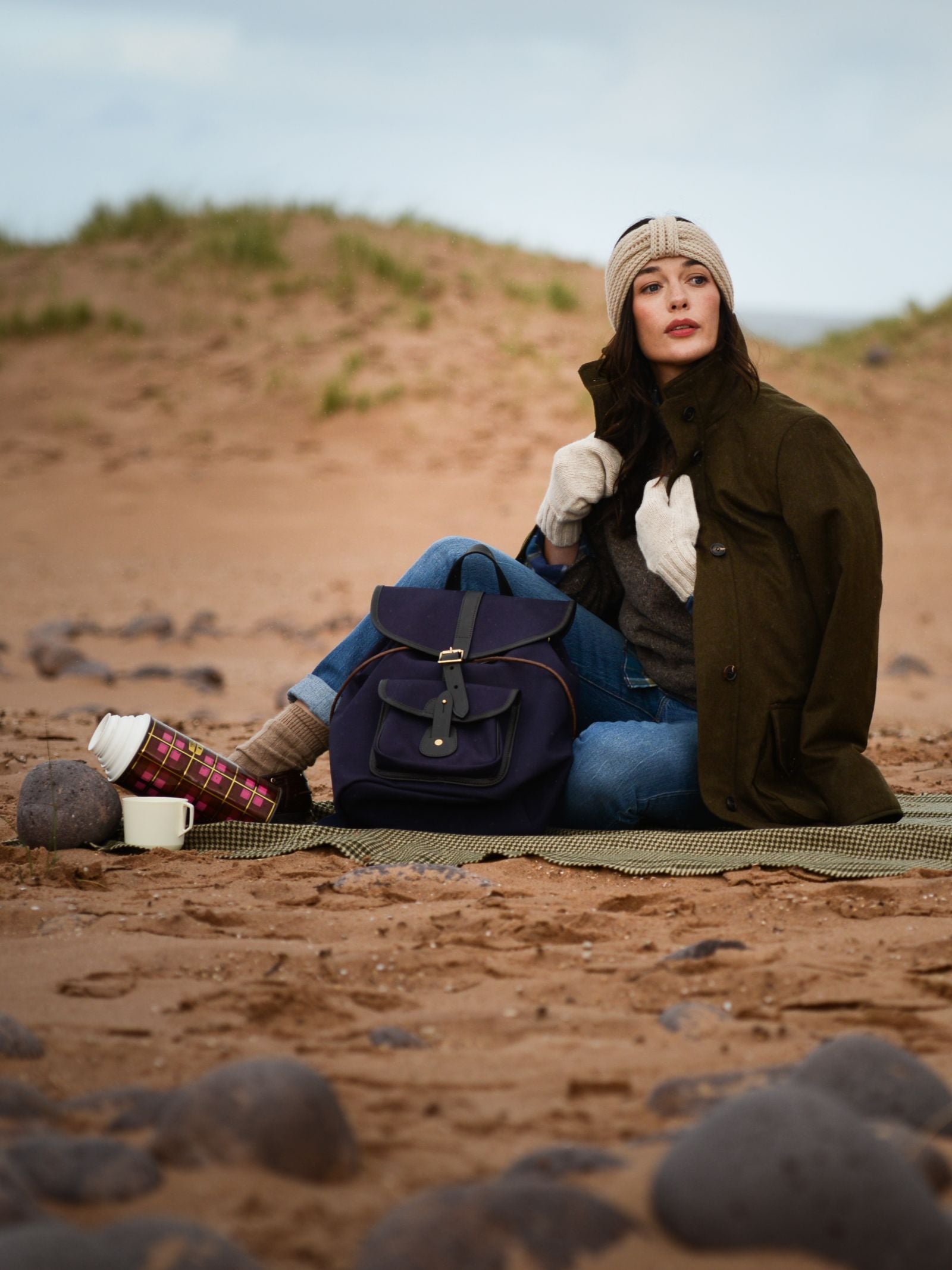 A woman sits on a blanket at a sandy beach, wearing a beanie, coat, jeans, boots, and Campbell's of Beauly Chunky Mittens. A blue backpack is by her side and a thermos with a mug rests among scattered rocks.