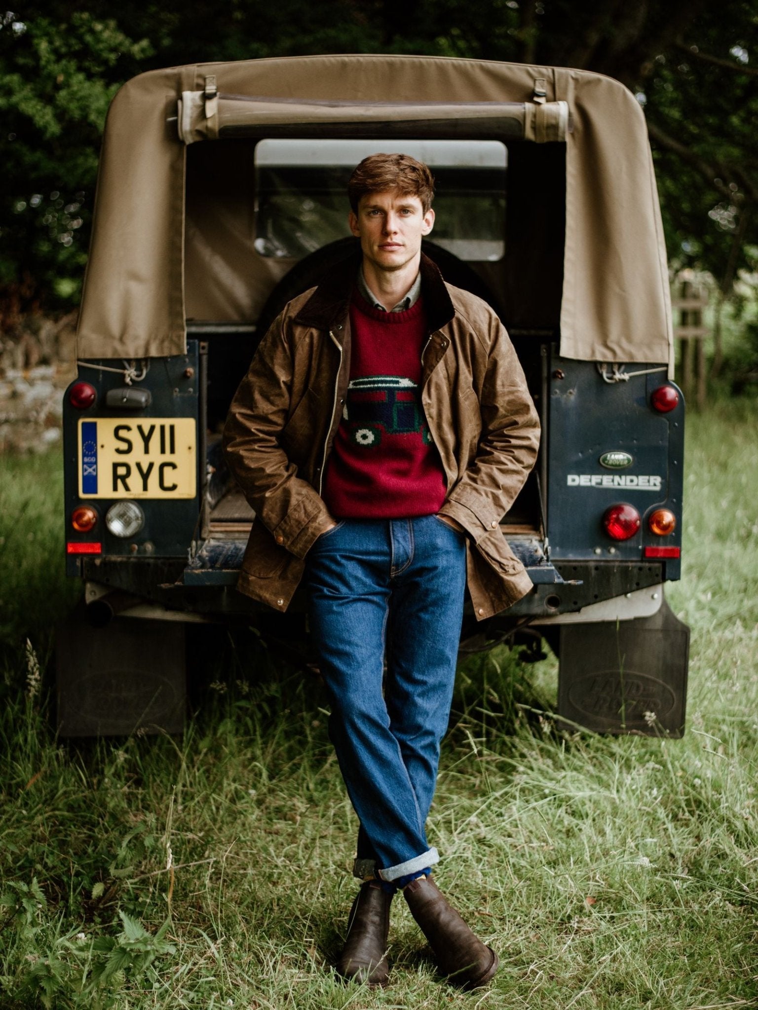 Wearing the Rain Cabaan by Campbell’s of Beauly—a classic brown waxed cotton jacket—a person in a red sweater and blue jeans leans against a tan Land Rover Defender parked on grass. Trees and greenery in the background evoke timeless heritage outerwear style.