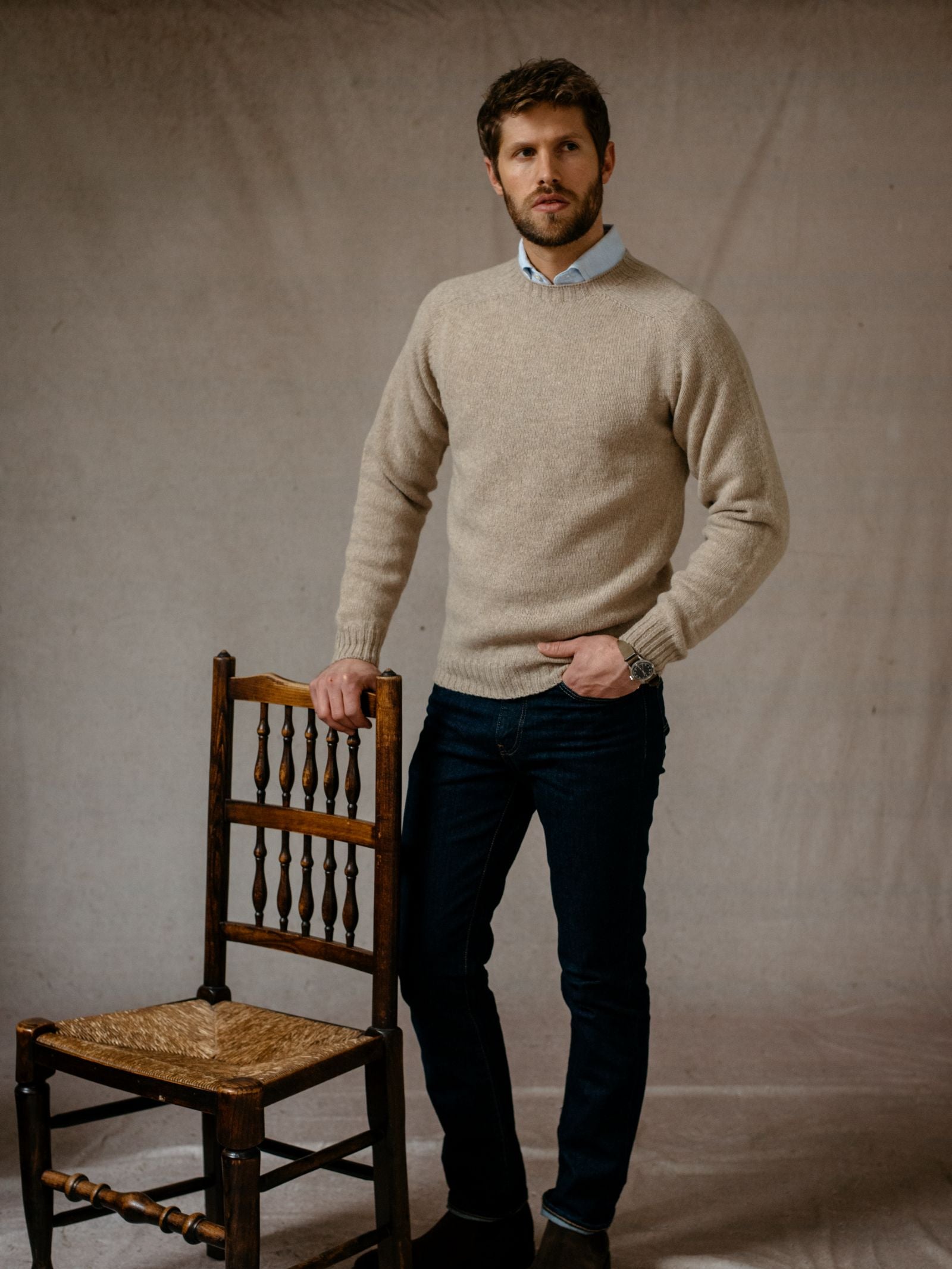 A man with short brown hair and a beard stands indoors beside a wooden chair, resting one hand on its back. He wears the Campbells of Beauly Shetland Jumper in heritage colourways, along with blue jeans and a collared shirt.