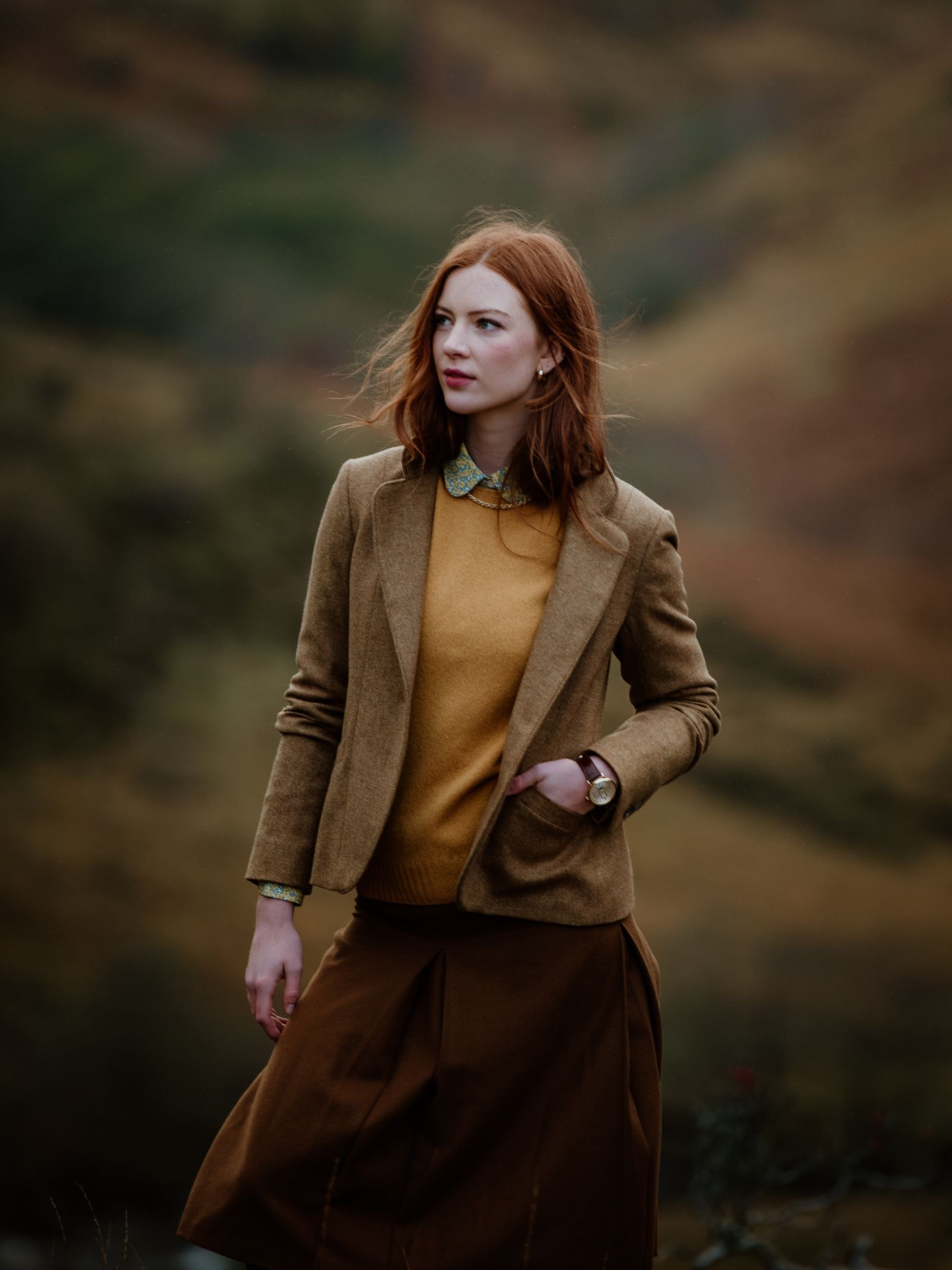 A woman with long red hair stands outdoors wearing the House Jacket by Campbells of Beauly, paired with a mustard sweater and brown skirt. A blurred natural background highlights her timeless style against the earthy landscape.