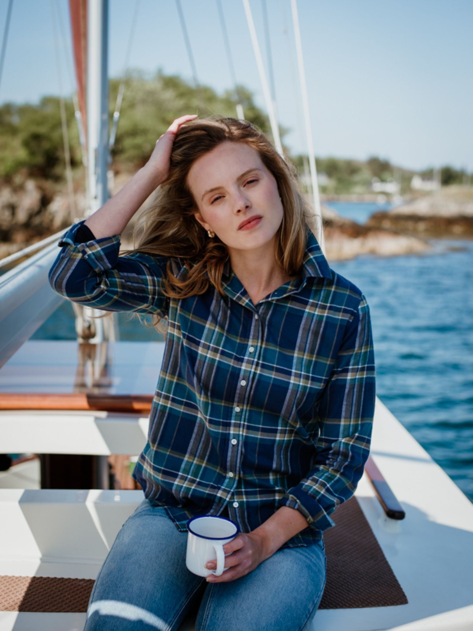 A woman wearing a blue Campbells of Beauly Brushed Cotton Shirt and jeans sits on a boat, holding a white mug and touching her hair, with sunny water and trees in the background.