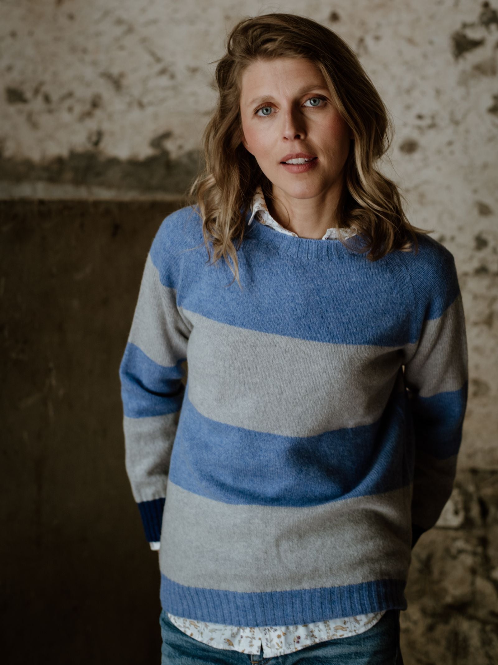 A woman with wavy blonde hair wears the Campbells of Beauly Stripe Crew Jumper, a blue and gray striped crew neck knit made in Scotland from premium yarns, as she stands before a weathered wall, looking at the camera neutrally.