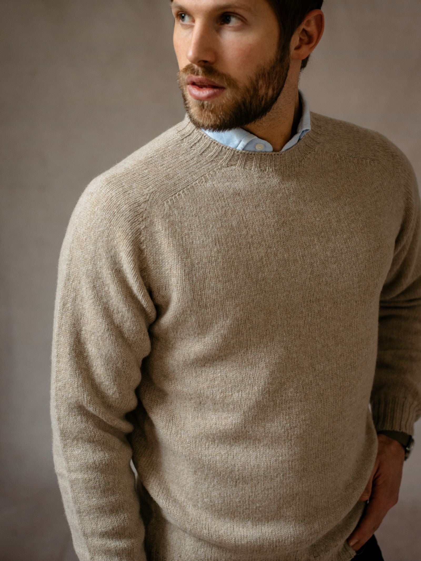 A bearded man stands against a neutral background, wearing a light brown Campbells of Beauly Shetland Jumper made from soft lambswool over a collared shirt, looking to the side with one hand in his pocket.