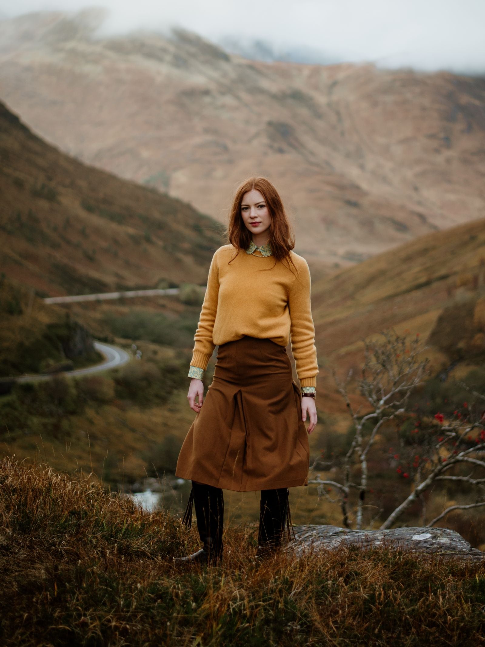 A red-haired woman stands on grassy terrain in the Scottish hills, wearing a Campbells of Beauly Geelong Lambswool Crew Neck Jumper over a collared shirt and brown skirt, with misty mountains and a winding road behind her.
