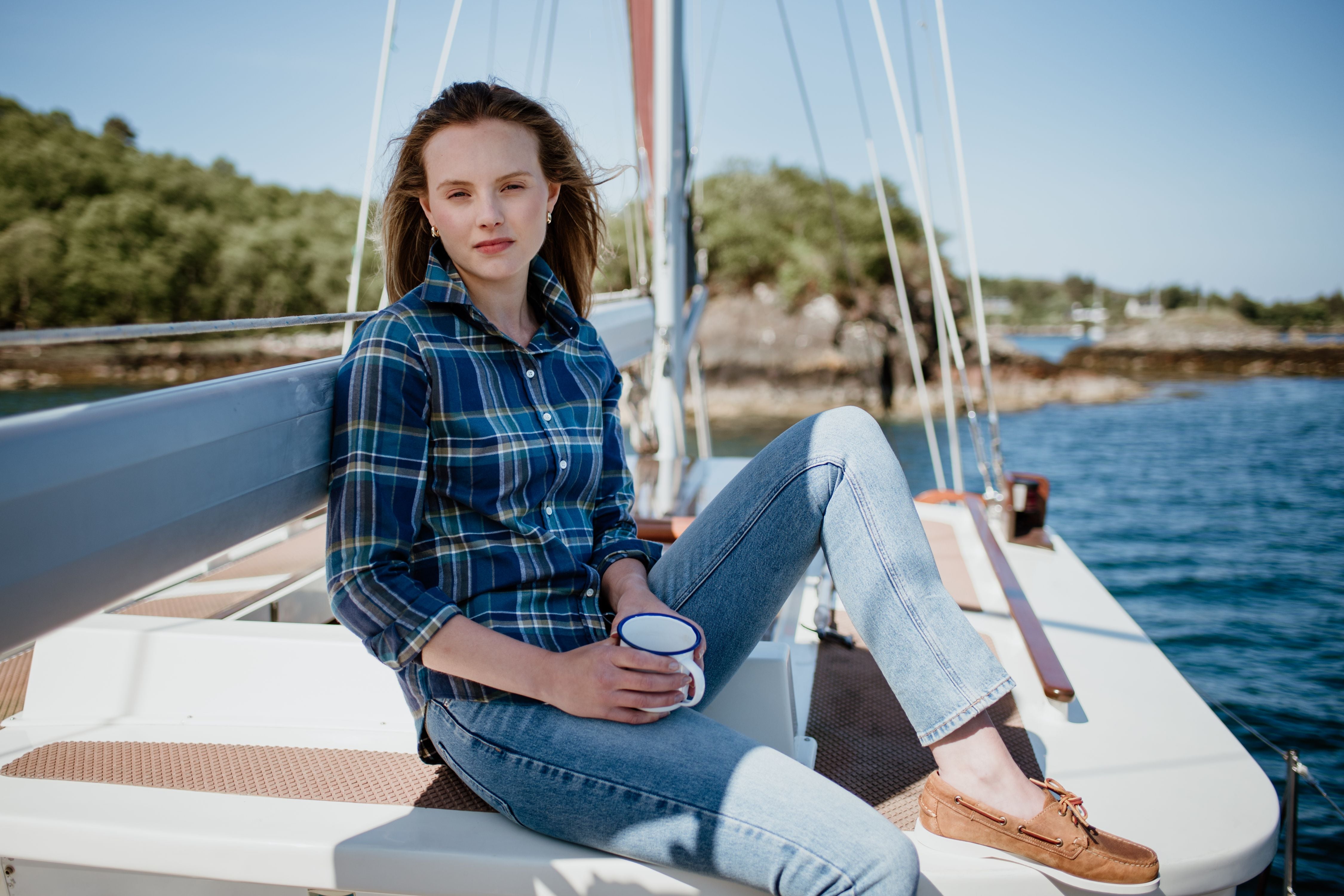 A young woman in a plaid shirt and jeans sits on the deck of a sailboat, holding a mug. Trees and rocky shoreline are visible in the background under a clear blue sky.