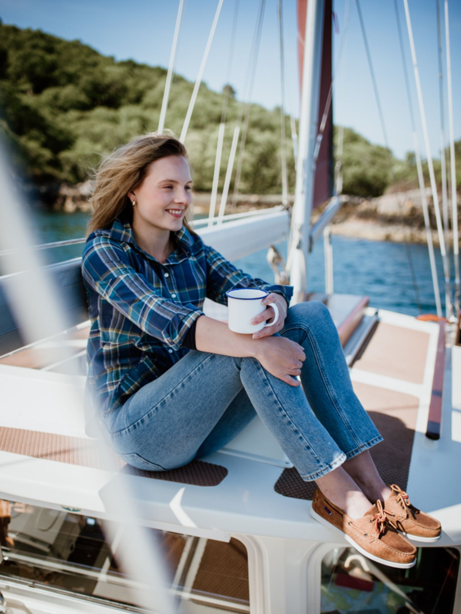 A woman in the Campbells of Beauly Brushed Cotton Shirt and jeans sits on a sailboat deck, holding a white mug and smiling by the water—showcasing this timeless, versatile wardrobe staple.