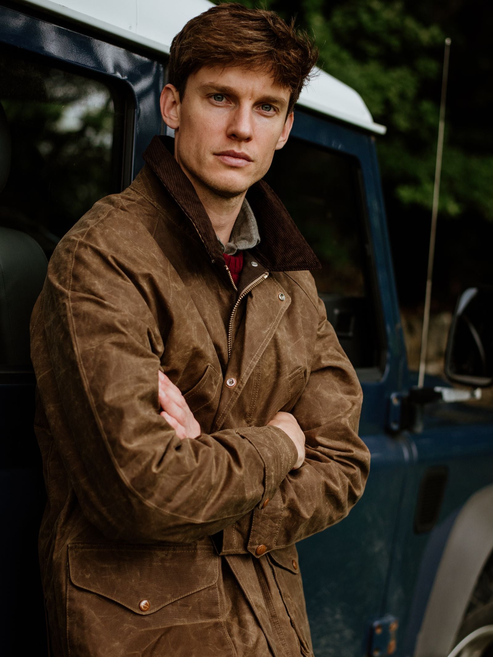 A man with short brown hair stands with arms crossed in front of a dark blue vehicle, wearing the Campbell's of Beauly Rain Cabaan Jacket—a classic brown waxed cotton coat. Green foliage appears in the background.