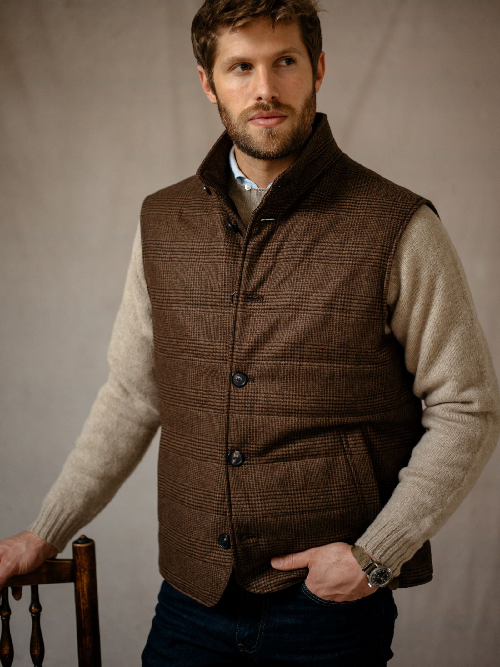 A man with short brown hair and a beard wears the Campbells of Beauly Cashmere Gun Club Quilted Gilet over a beige sweater, dark jeans, and a wristwatch. He stands by a wooden chair, gazing to the side against a neutral background.
