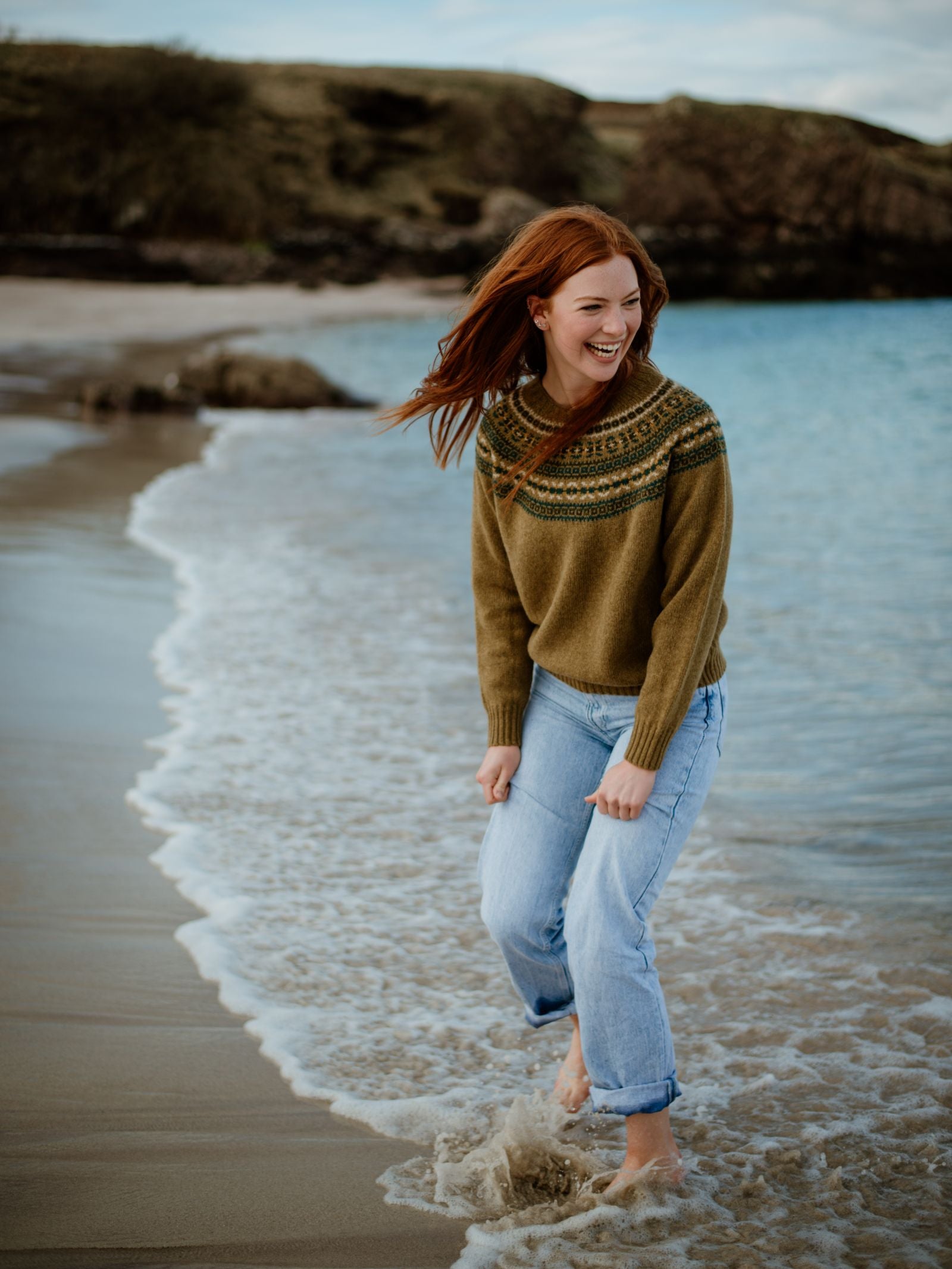 A woman with long red hair, wearing the Campbells of Beauly Fairisle Crew Jumper and rolled-up jeans, smiles as she walks barefoot along the shoreline with gentle waves, set against a backdrop of rocky cliffs and calm water.