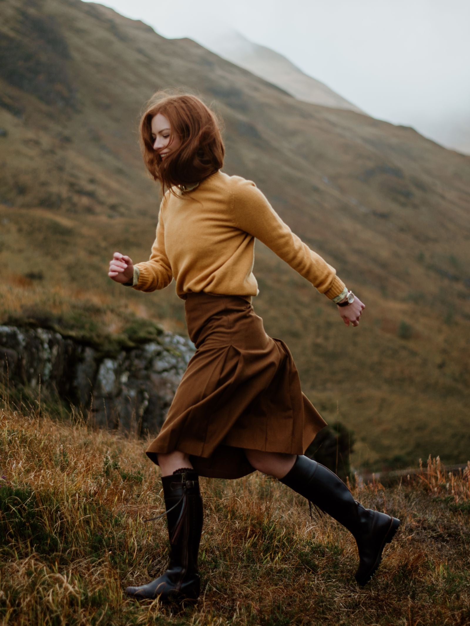 A woman with reddish hair, wearing a mustard sweater, brown skirt, and Campbells of Beauly Leather Spanish Boots, walks briskly through grassy hills with mountains in the background on a cloudy day.