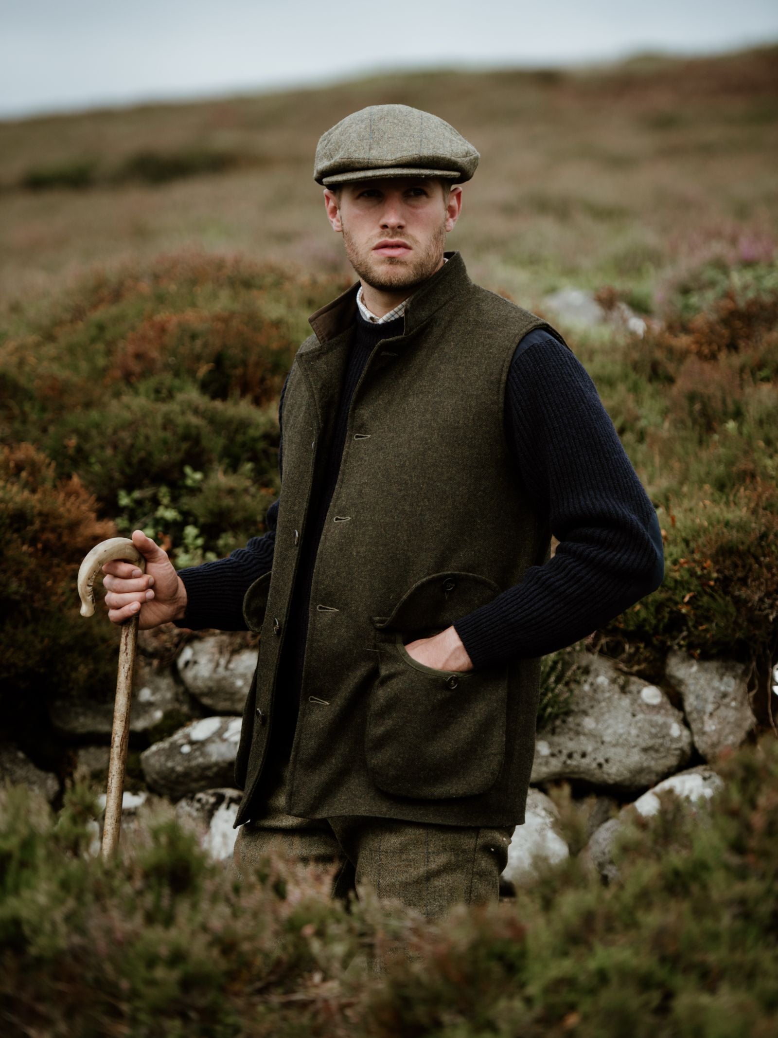 A man in a Campbells of Beauly Field Nehru Gilet with quilted lining, flat cap, and sweater stands on grassy hills with a wooden walking stick, gazing slightly away from the camera.