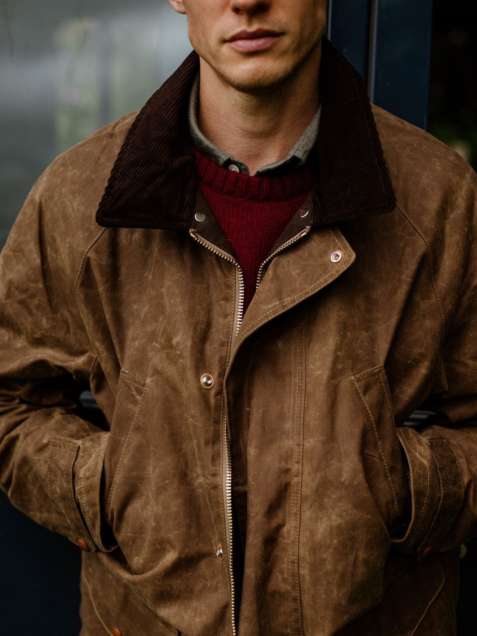 A man stands with hands in pockets, wearing the Rain Cabaan—a brown waxed cotton jacket by Campbell’s of Beauly, featuring a dark collar—layered over a red sweater and collared shirt; his face is partially visible, embodying classic heritage outerwear.