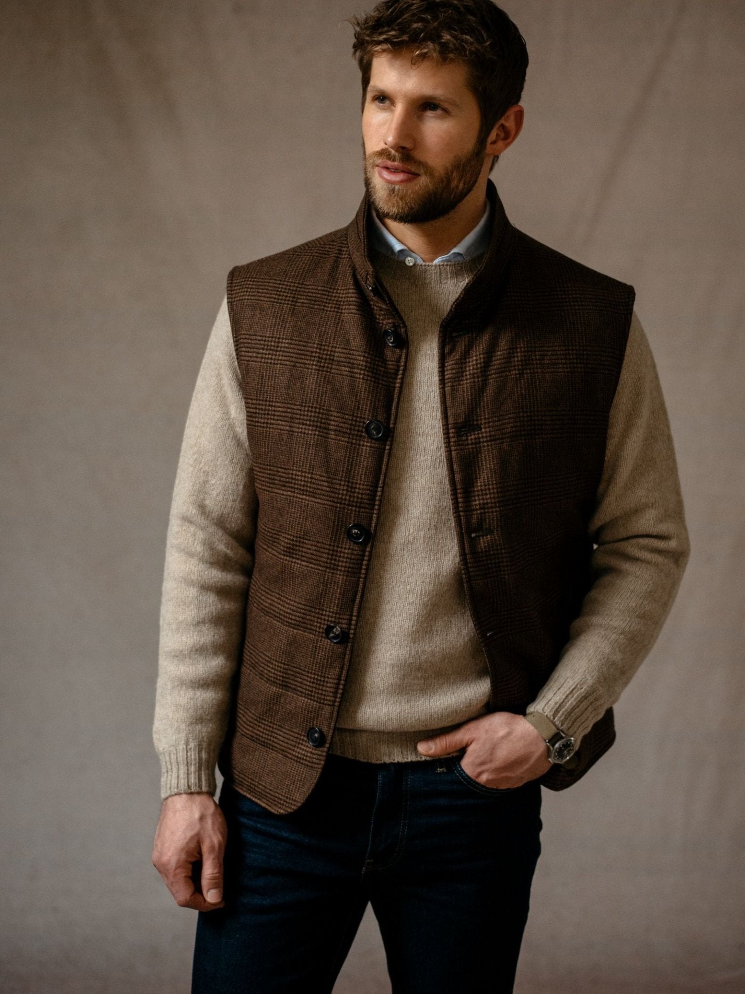 A man wears the Campbells of Beauly Cashmere Gun Club Quilted Gilet over a beige sweater with dark jeans, standing against a neutral background and looking to the side with one hand in his pocket.