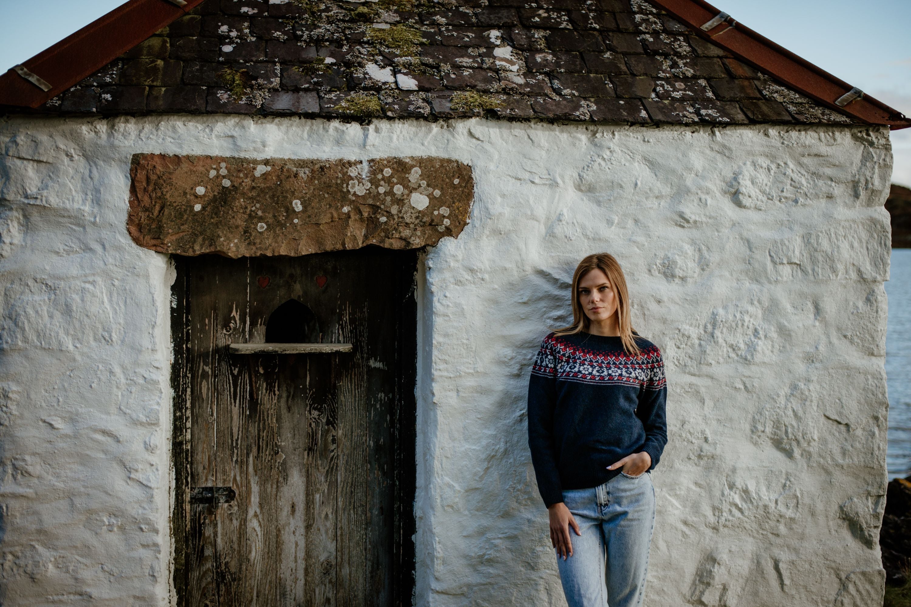 A woman wearing a dark blue sweater with a red and white pattern and jeans stands by a rustic whitewashed stone building with a weathered wooden door and a mossy roof.