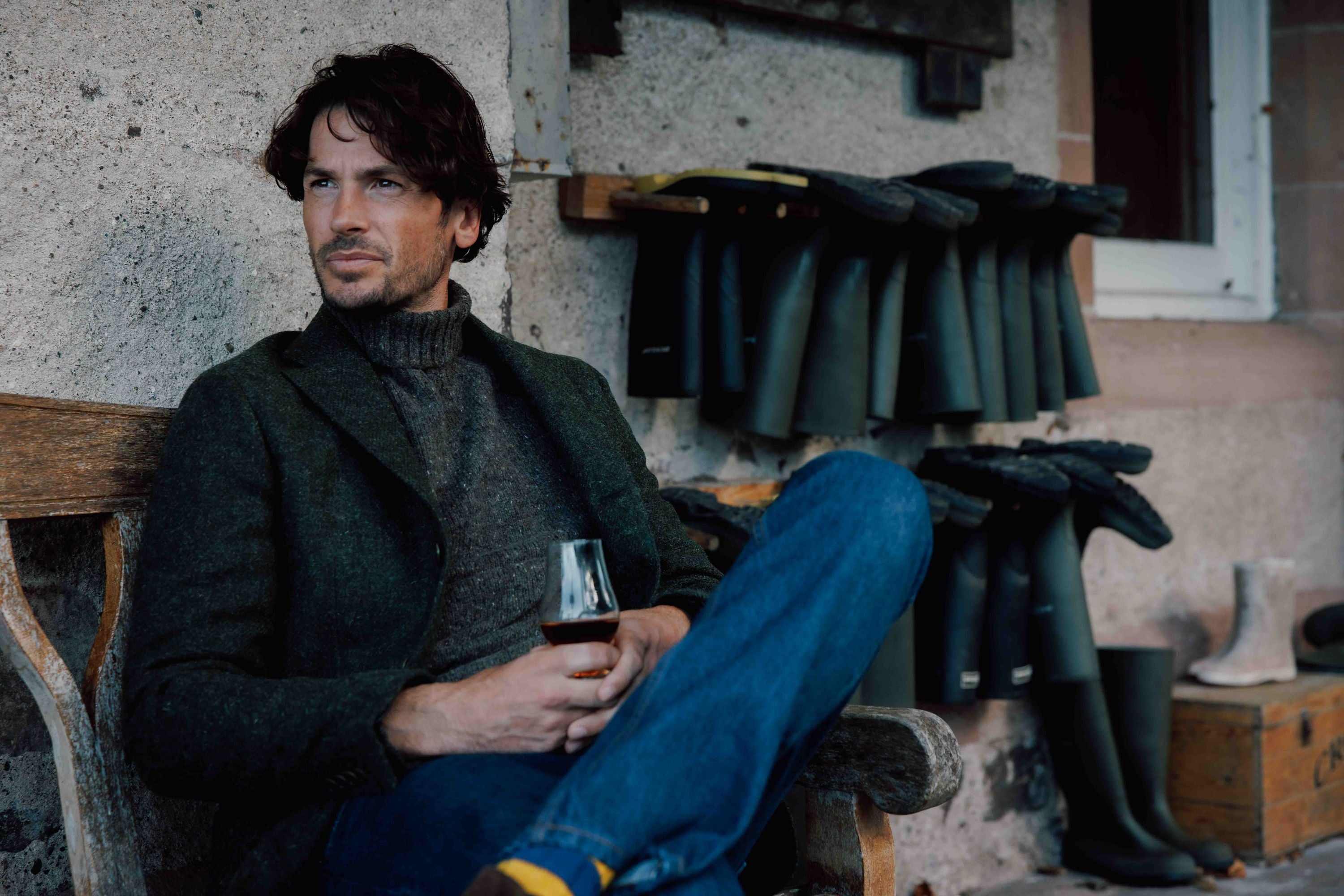 A man with brown hair sits on a wooden bench, holding a glass of wine. He wears a dark jacket, gray turtleneck, and jeans. Behind him, several pairs of green rubber boots are lined up against a wall.