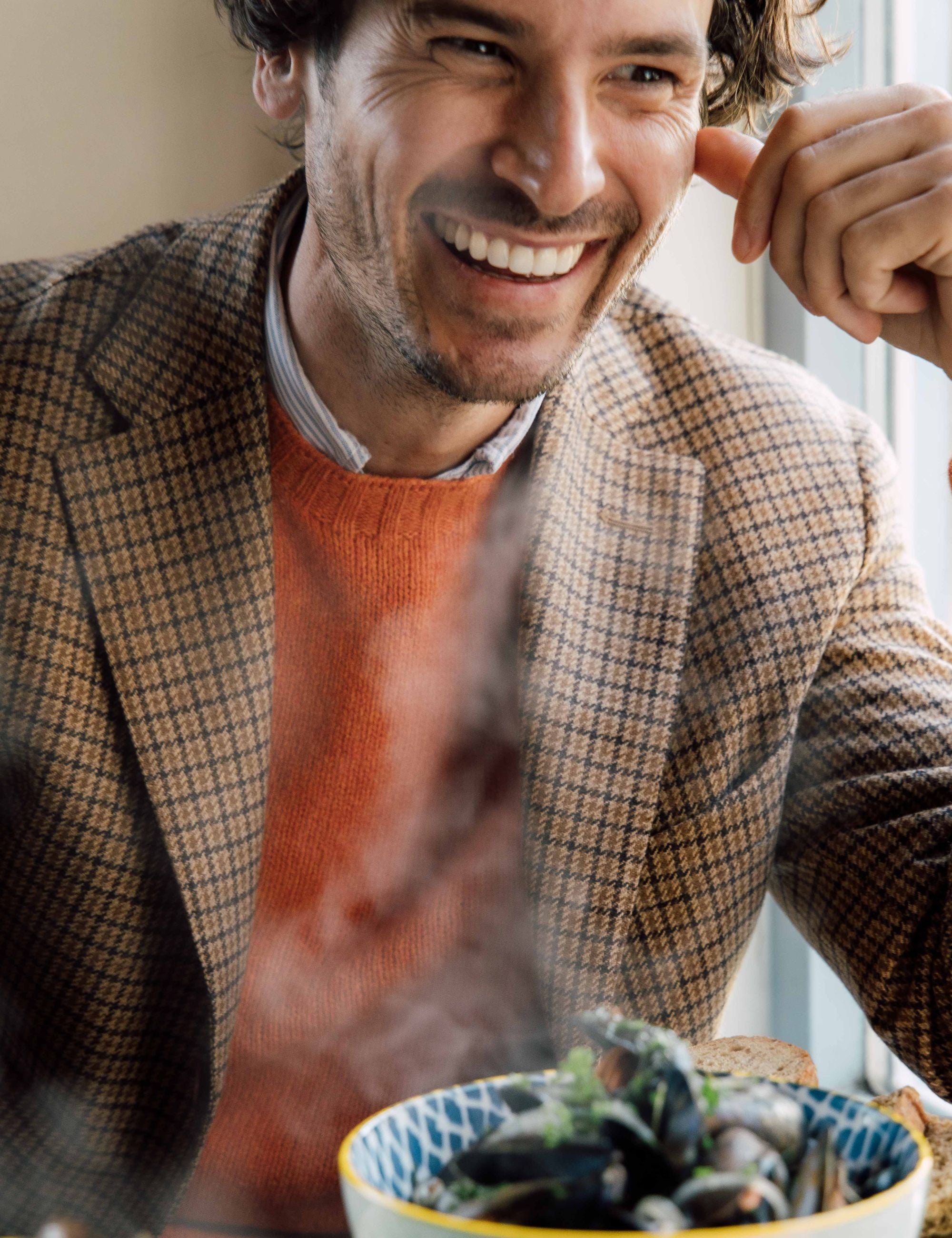 A smiling man in a brown plaid blazer and orange sweater sits at a table with a steaming bowl of food in front of him, enjoying a meal in a bright, cozy setting.