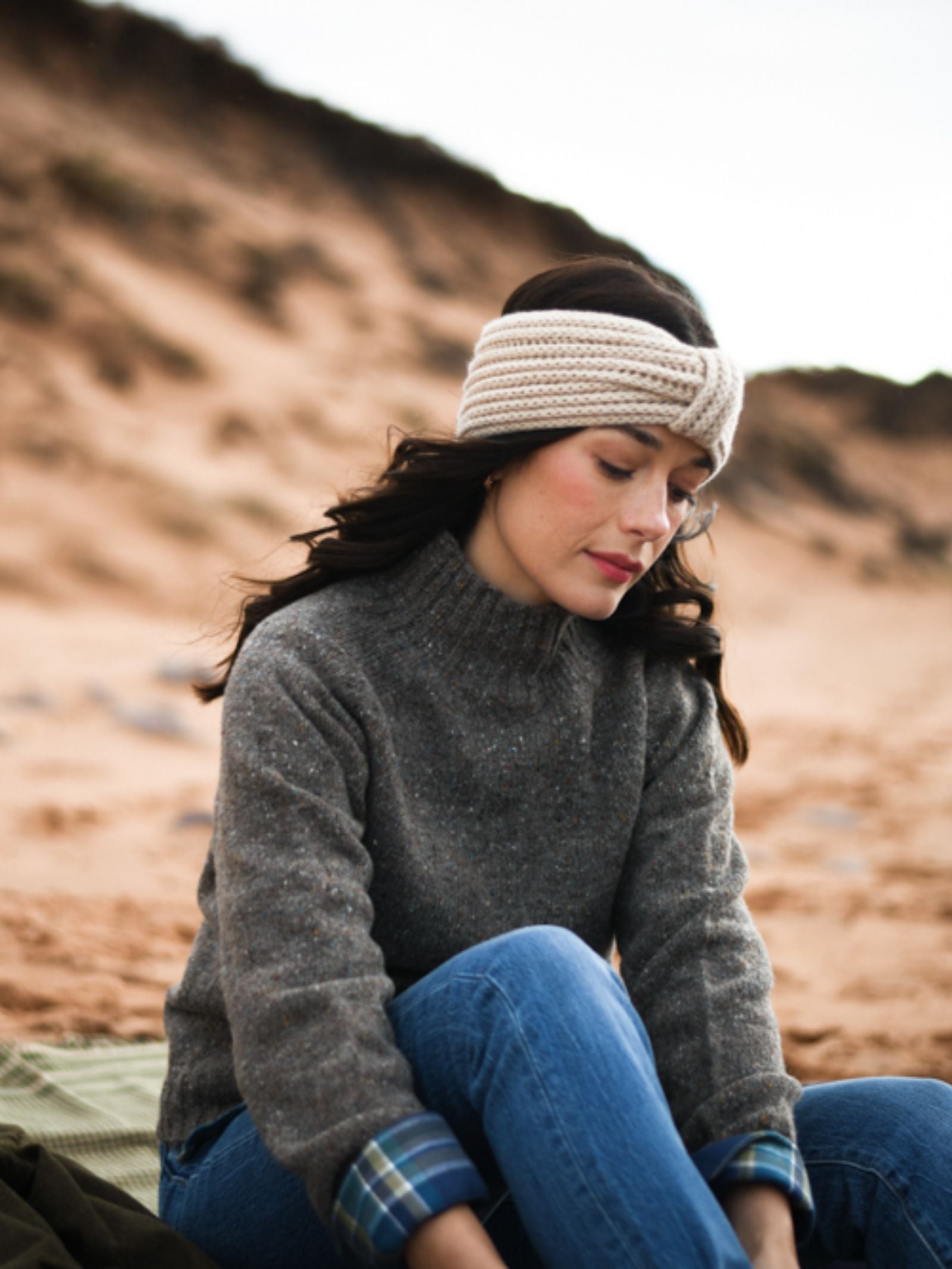 A woman with long dark hair wears the Campbell's of Beauly Donegal Lambswool Funnel Neck Jumper, blue jeans, and a beige knit headband as she sits thoughtfully on a sandy beach with cliffs in the background.