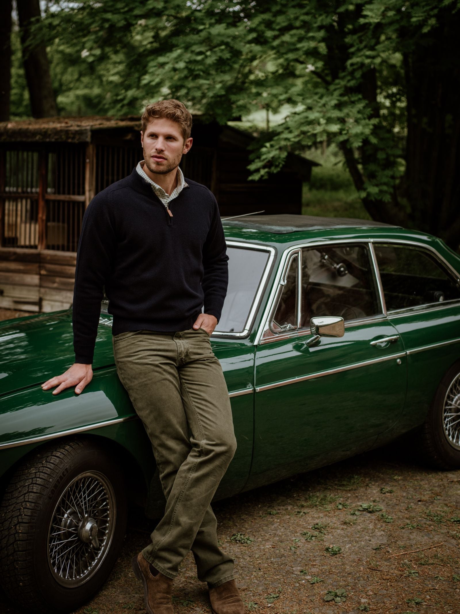 A man with short brown hair and a beard leans against a classic green car outdoors, wearing Campbells of Beauly Lambswool Quarter Zip Jumper, olive pants, and brown boots amid trees and a rustic wooden structure in the Scottish Borders.