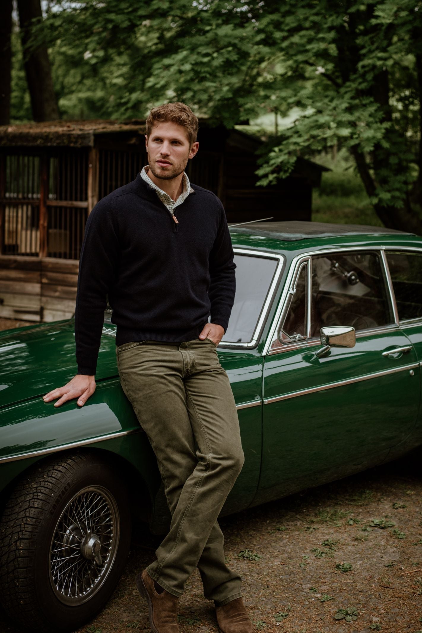 A man in a Campbells of Beauly Lambswool Quarter Zip Jumper and olive pants leans against a classic green car parked outdoors, with trees and wooden structures in the background.