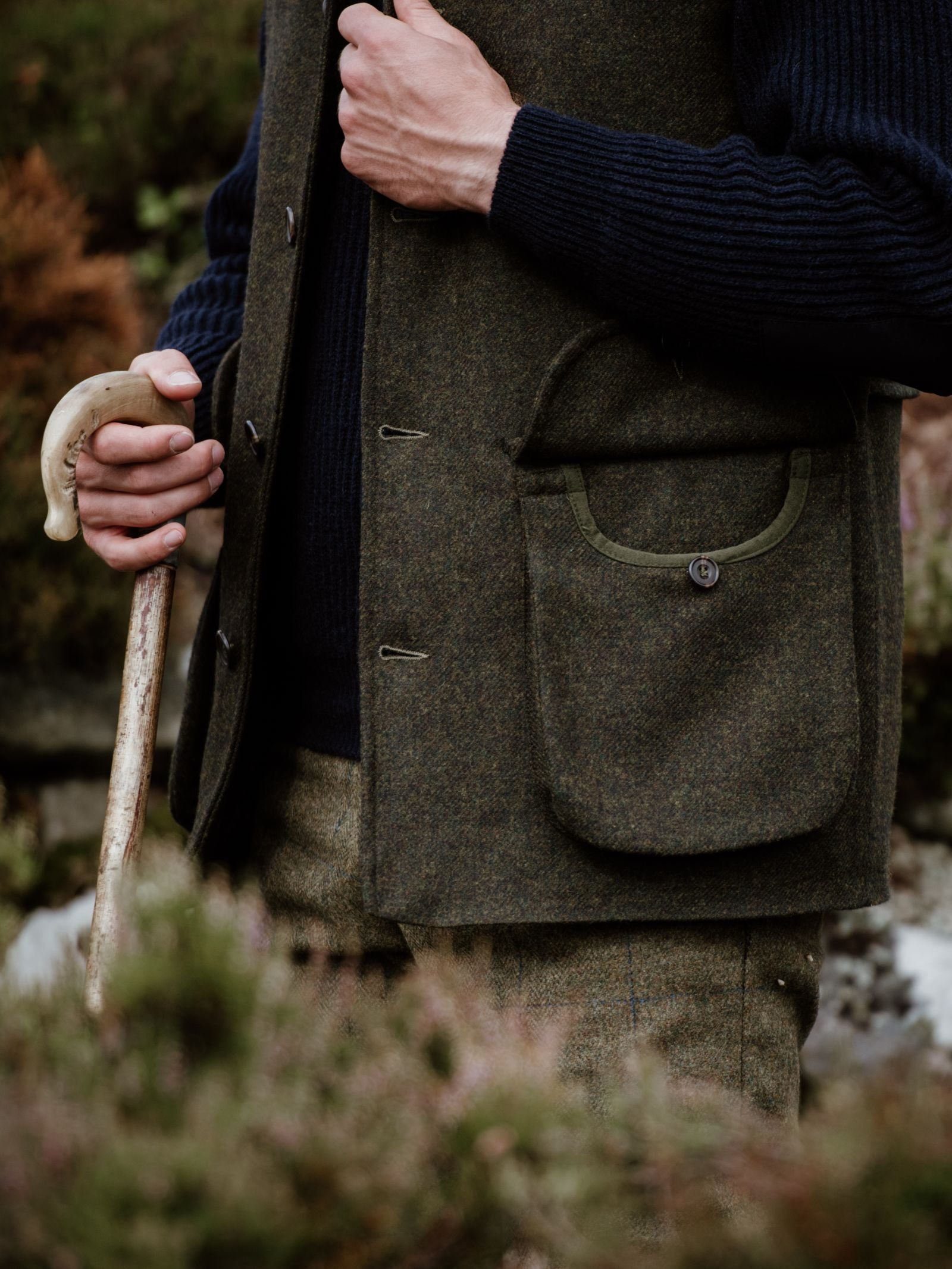 Wearing the Campbells of Beauly Field Nehru Gilet in olive-green moleskin over a navy ribbed sweater and matching trousers, a person stands outside holding a wooden cane, with one hand near the gilet pocket amid blurred plants and rocks.