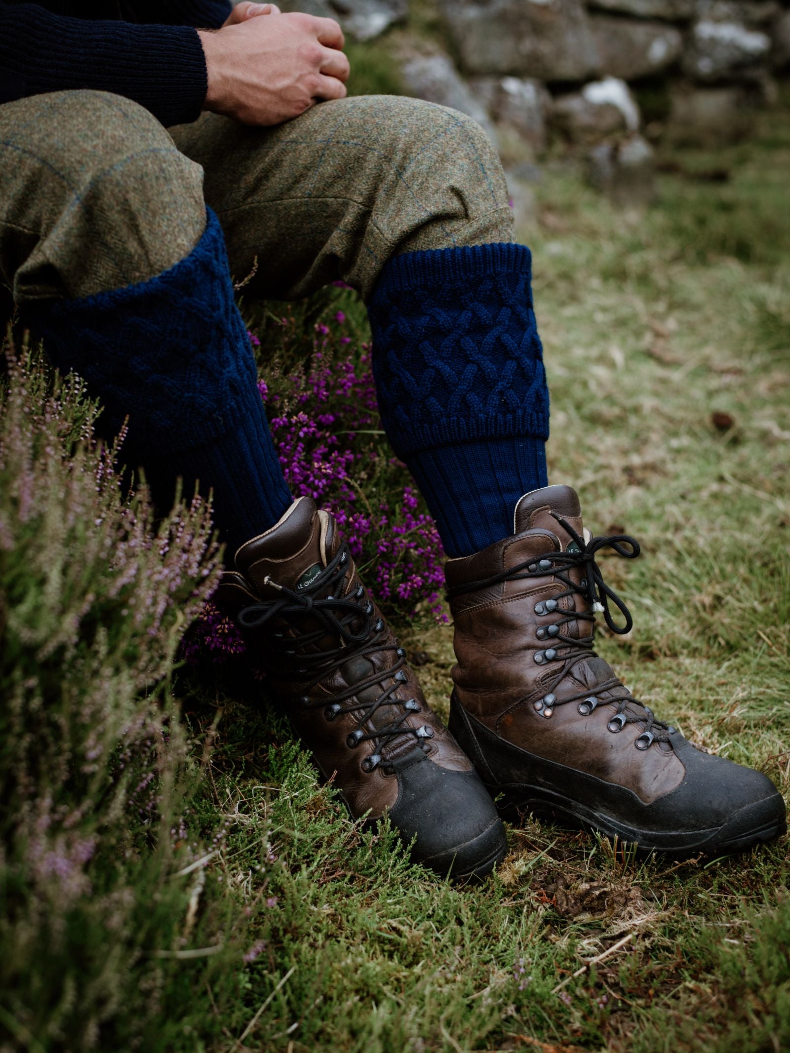 A person sits outdoors on grass, wearing Campbells of Beauly Merino Rannoch Shooting Socks in a blue pattern with brown hiking boots and rolled-up tweed trousers. Purple heather and rocks appear in the background.