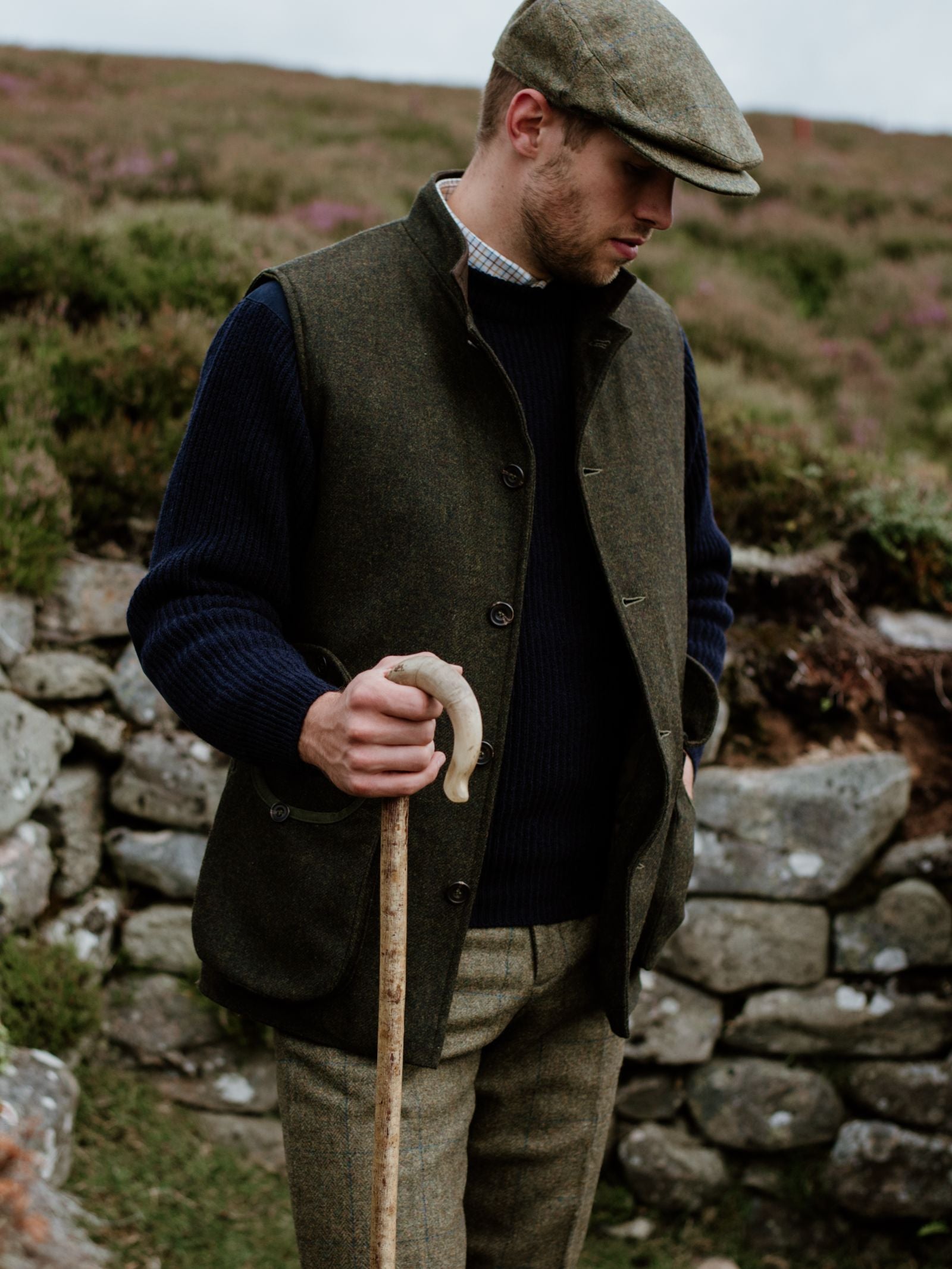 A man wearing Campbells of Beauly Plus Twos—classic country attire with a flat cap, vest, and trousers—stands outdoors by a stone wall with a wooden walking stick. The grassy background is softly blurred.