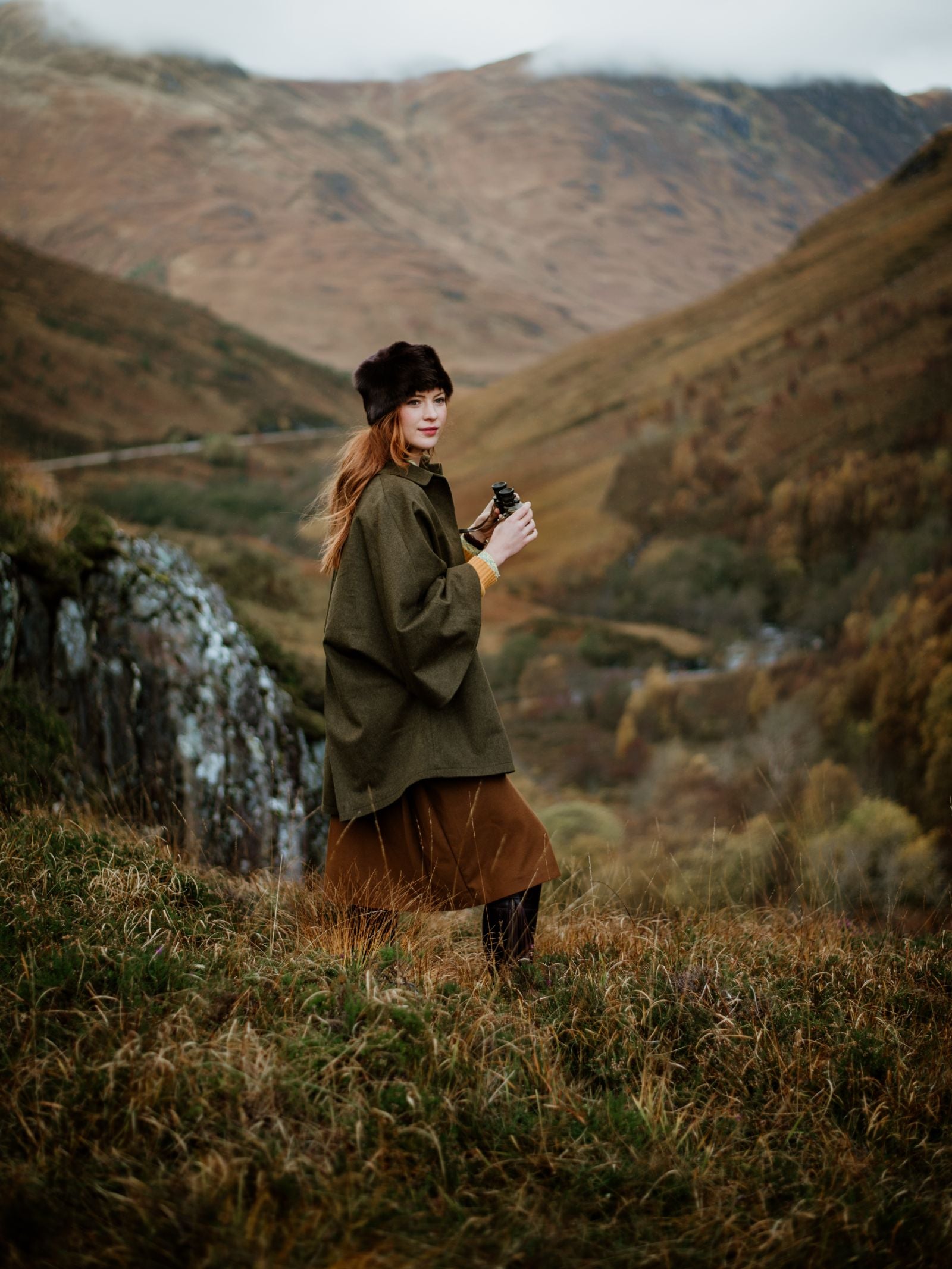 A woman with long hair stands in a grassy valley, holding binoculars and wearing the Campbells of Beauly Loden Green Cape. Hills and mountains with autumn colors and cloudy skies form the scenic background.