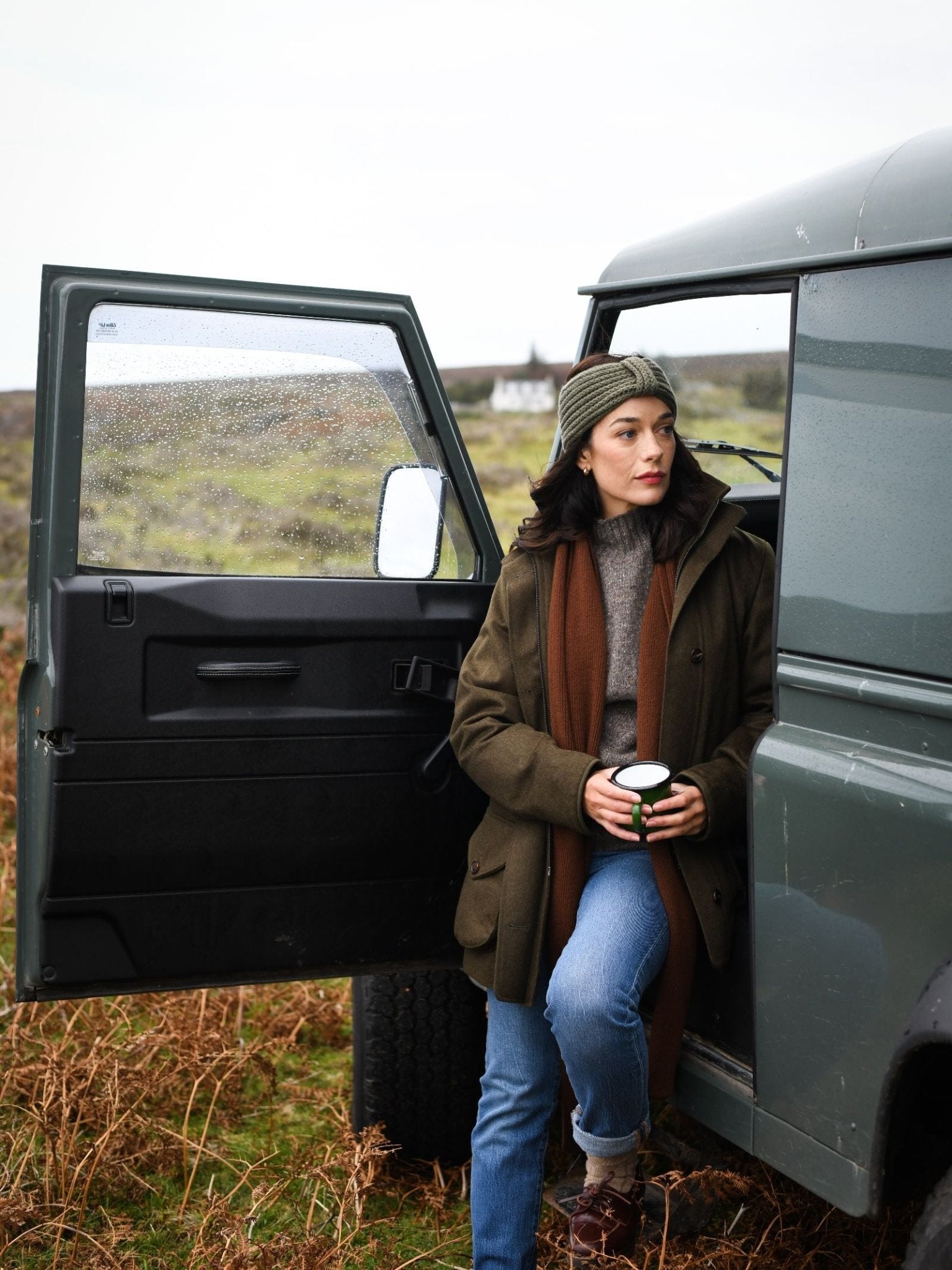 A woman wearing a Campbell's of Beauly Cashmere Ear Warmer leans against a green SUV with the door open, holding a mug. She stands among grass and ferns, enjoying her cozy accessory on a cloudy countryside day.